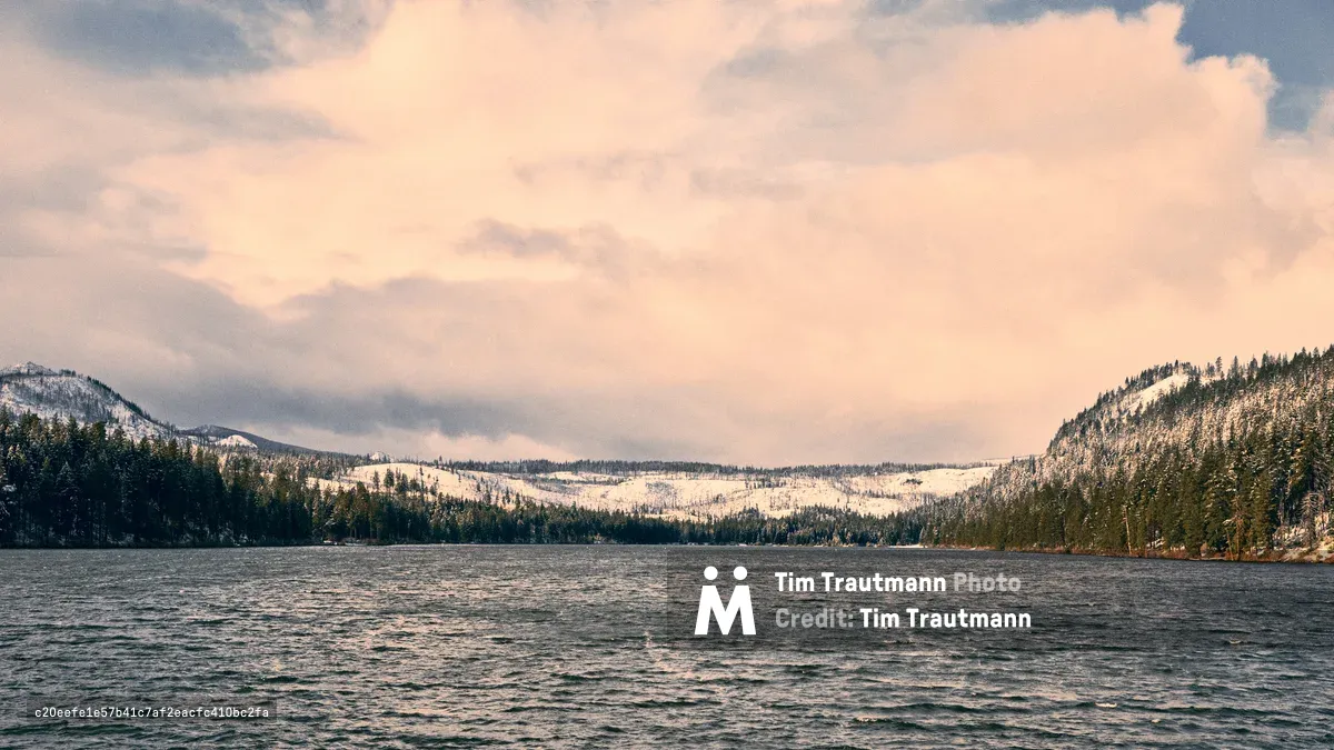 Dusky rose and lavender clouds drift across the evening sky above Oregon's Suttle Lake, where dark waters ripple gently between snow-dusted Cascade foothills. The serene alpine lake reflects the soft pastel light of approaching winter dusk, while dense coniferous forests cloak the surrounding mountains in deep green shadows. This tranquil moment captures the pristine wilderness of Jefferson County's high country, where the lake's surface mirrors the ephemeral beauty of the mountain sunset.