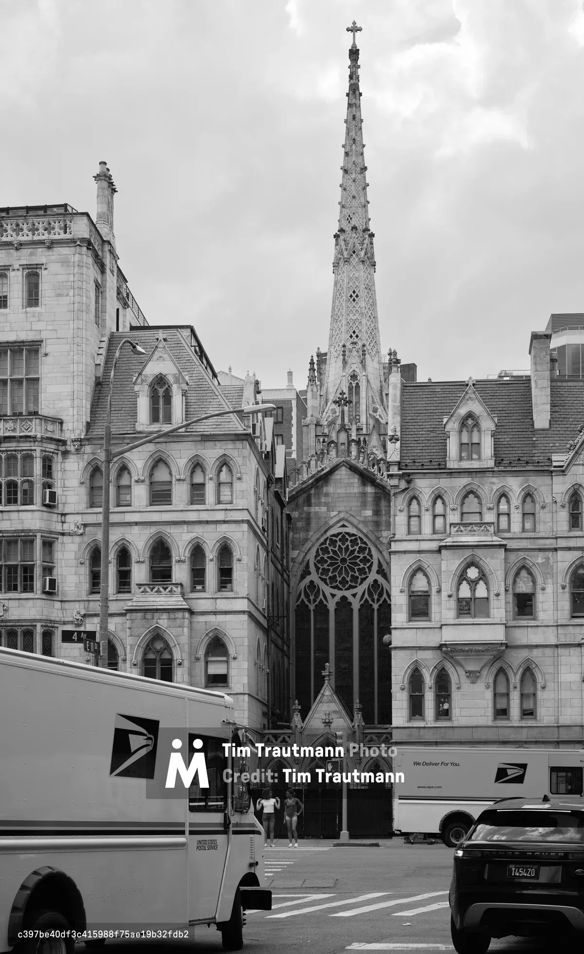 Grace Church's ornate Gothic Revival spire soars majestically above the bustling streets of Greenwich Village, its intricate stonework and cross-topped pinnacle creating a dramatic vertical counterpoint to the surrounding historic buildings. The black and white composition emphasizes the architectural details of the church's elaborate facade, complete with rose window and pointed arches, while postal trucks and urban activity ground the sacred structure in contemporary New York life. Soft, overcast lighting bathes the limestone surfaces in gentle gradations of gray, highlighting the craftsmanship of this 1840s ecclesiastical masterpiece.