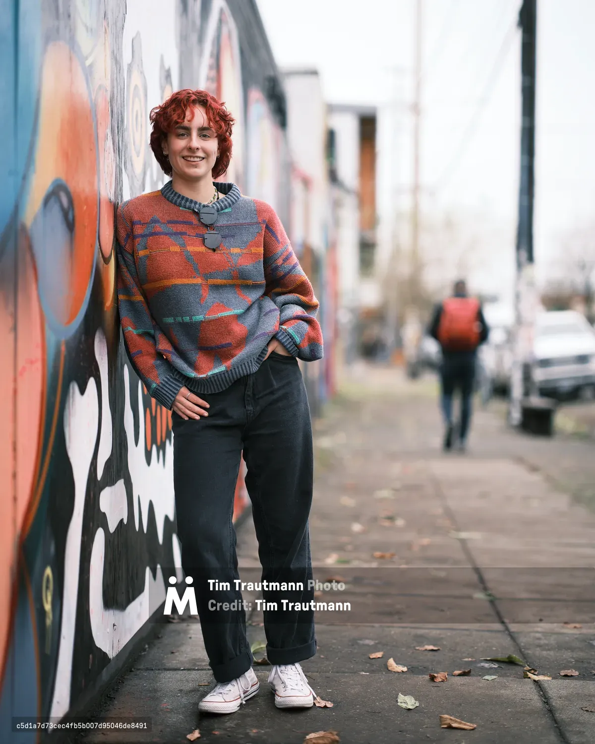 A full-length senior portrait of Annika Molina leaning against a colorful street mural on a Portland sidewalk. She has short curly red hair and wears an oversized patterned sweater in orange, grey, and teal, dark wide-leg jeans cuffed at the ankle, white Converse sneakers, and a pendant necklace. She smiles warmly at the camera with one hand on her hip. A pedestrian with a red backpack walks in the blurred background along the tree-lined street.