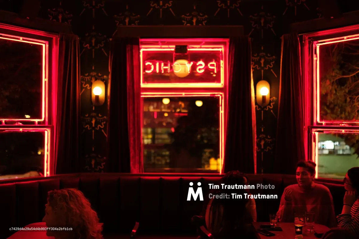 The iconic neon-framed windows of Psychic Bar on North Mississippi Avenue bathe patrons in an otherworldly crimson glow. The reversed "PSYCHIC" sign blazes against ornate wallpaper while warm amber street lights create depth beyond the glass. Silhouetted figures occupy red-washed tables in the foreground, their forms dissolved into the moody atmospheric lighting that defines this beloved Portland establishment.