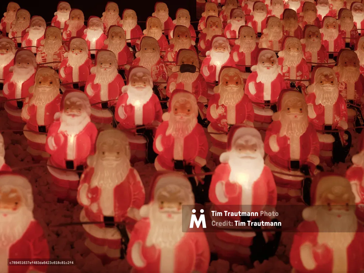 Dozens of identical illuminated Santa Claus figurines create a mesmerizing sea of red and white in Portland's Sellwood neighborhood. The glowing plastic figures, part of Chris Willis's annual art installation, cast warm amber light against the darkness, their repetitive forms suggesting both festive abundance and unsettling uniformity. Each Santa bears the same benevolent expression and raised hand gesture, creating a hypnotic pattern that stretches beyond the frame's edges.