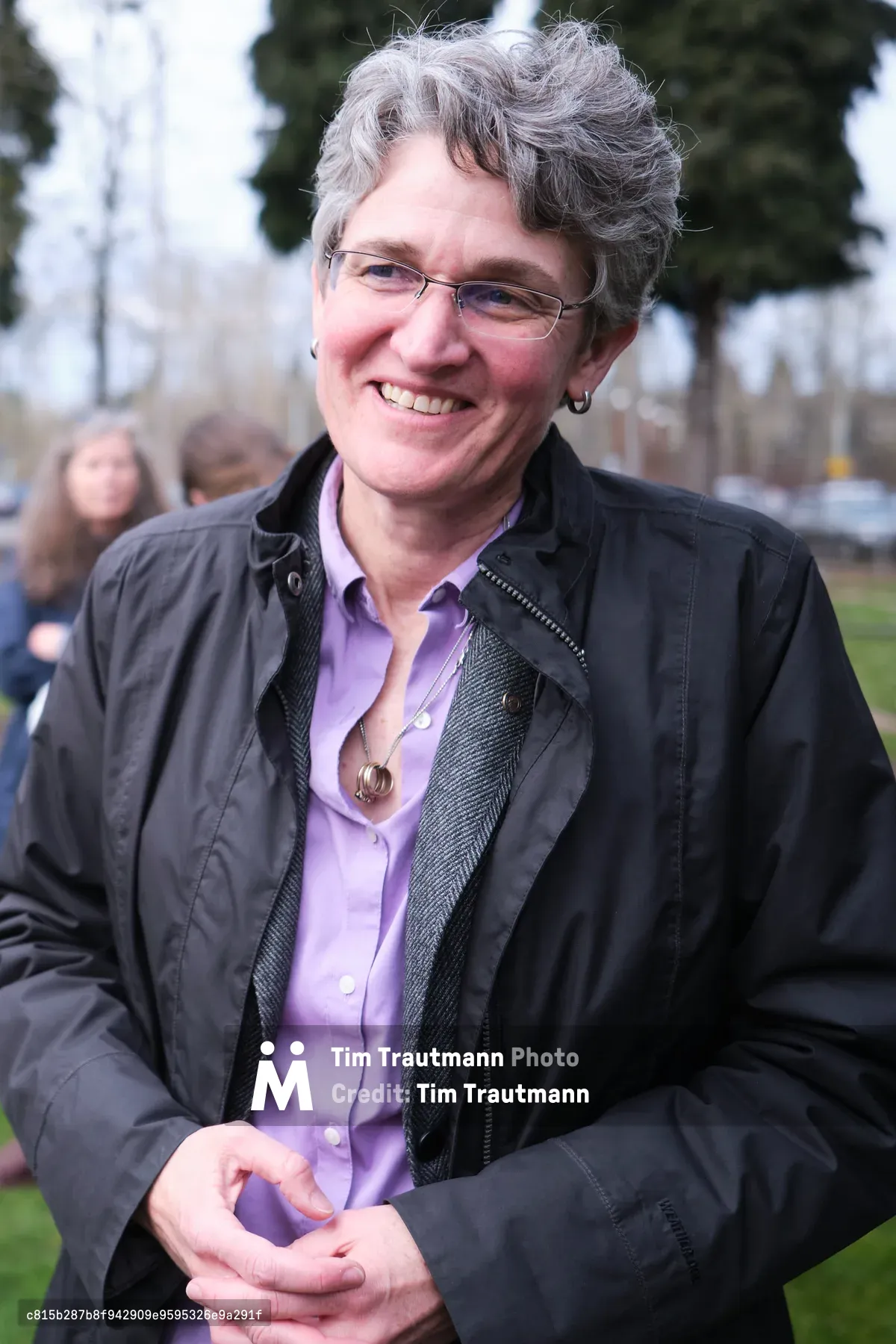 A middle-aged woman with curly gray hair and glasses smiles warmly at the camera while wearing a purple button-up shirt under a dark jacket. She has her hands clasped together and is photographed outdoors in what appears to be a park or community gathering space.