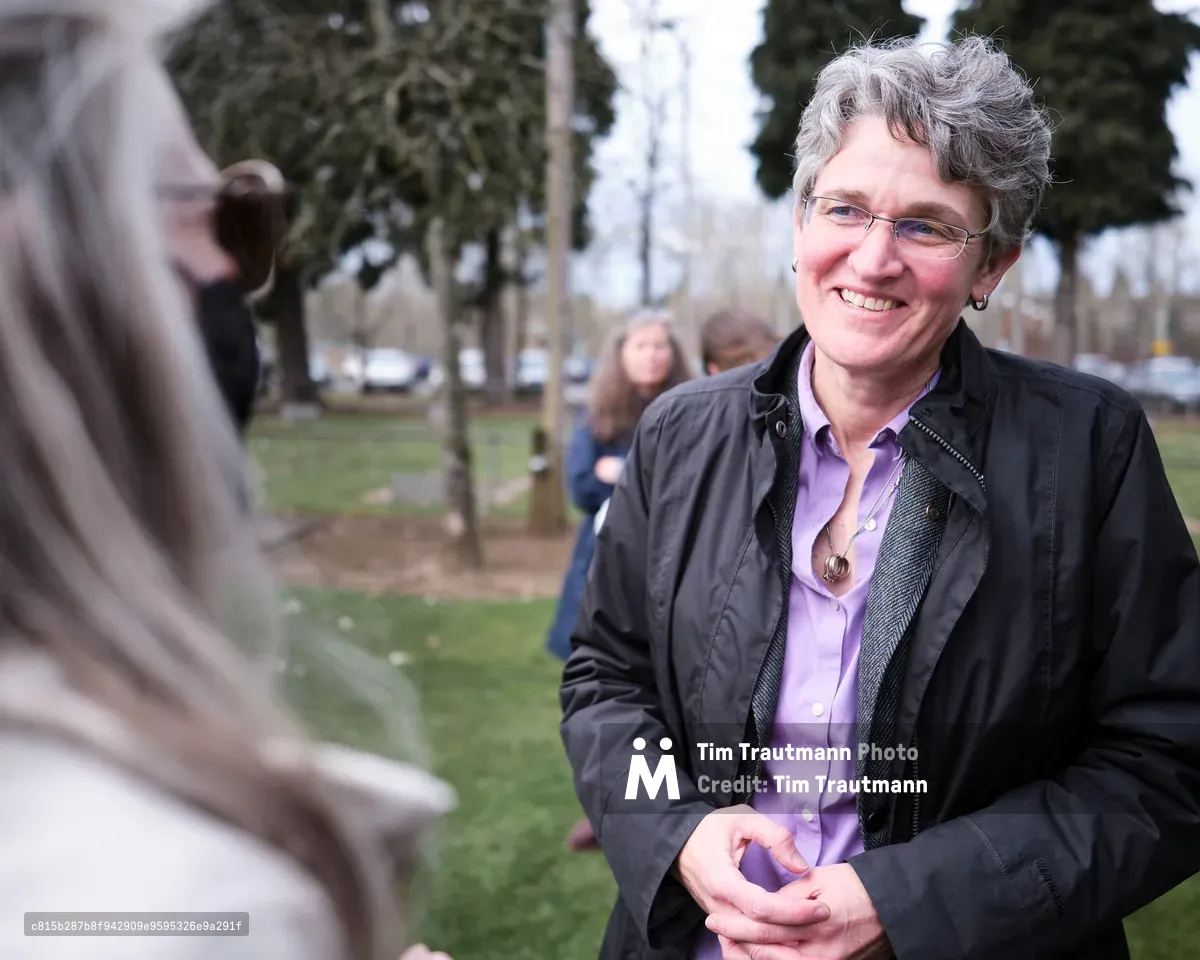 A middle-aged woman with gray curly hair and glasses smiles warmly while engaged in conversation in a park setting. She wears a black jacket over a purple shirt, standing in what appears to be a public space in Oregon City with trees and other people visible in the blurred background.
