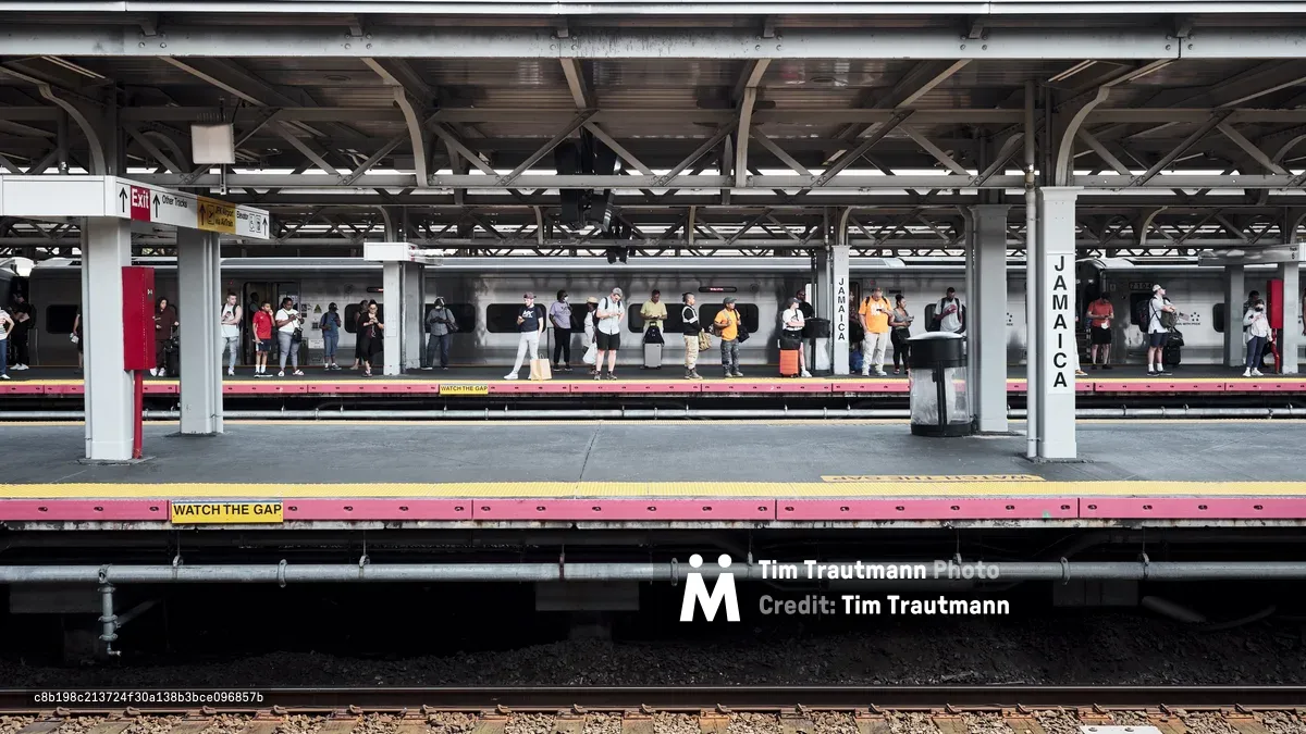 Passengers disperse across the weathered platform of Jamaica Station, their colorful summer clothing creating vibrant punctuation marks against the industrial backdrop of steel beams and concrete pillars. The AirTrain car sits silent between destinations, its sleek silver form contrasting with the utilitarian architecture of this crucial transportation hub. Harsh fluorescent lighting mingles with natural daylight filtering through the elevated structure, casting complex shadows that emphasize the geometric patterns of the overhead framework.