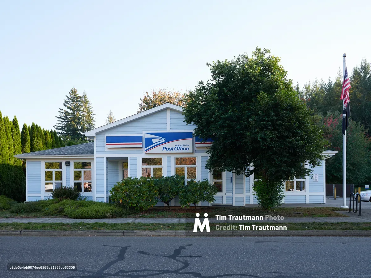 The modest United States Post Office in Amboy, Washington stands quietly beneath a pale evening sky, its white clapboard siding and iconic blue postal service signage creating a portrait of rural American infrastructure. Mature evergreens and a stately deciduous tree frame the single-story building, while an American flag stands sentinel at the property's edge. The soft, diffused light captures the unhurried pace of this small Clark County community, where the post office remains a vital civic anchor along Northeast 399th Street.