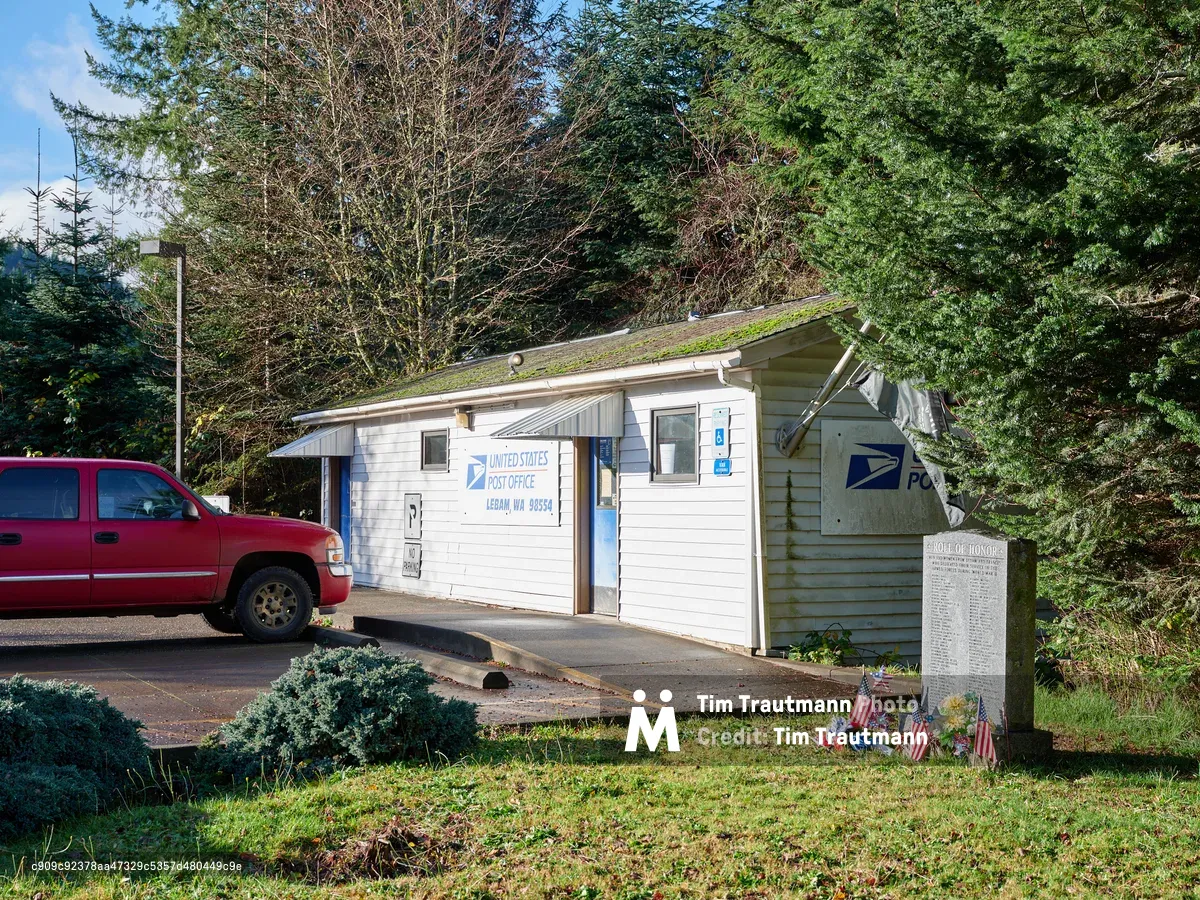 A weathered white clapboard building serves as the United States Post Office in the tiny community of Lebam, Washington. The modest single-story structure, crowned with a moss-covered roof that speaks to the Pacific Northwest's perpetual dampness, sits beneath towering evergreens that frame the scene. A red pickup truck idles nearby while small American flags planted near a memorial honor board create a poignant display of small-town patriotism. The late afternoon light filters through the forest canopy, casting a gentle glow on this quintessential slice of rural American postal service.