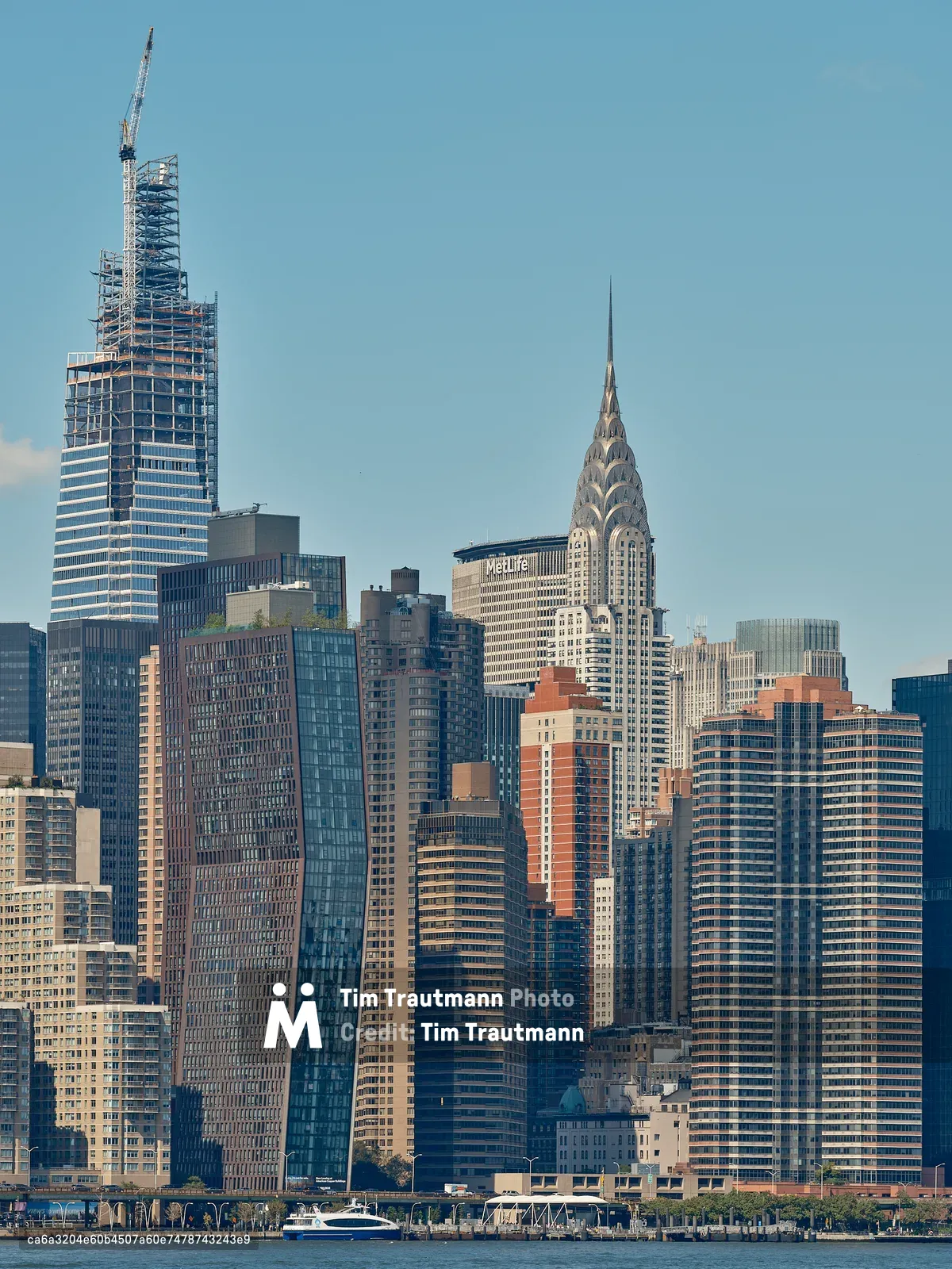 The Midtown Manhattan skyline viewed from across the East River under a clear blue sky. One Vanderbilt tower is visible under construction on the upper left, its steel frame topped with a construction crane. The iconic Art Deco Chrysler Building with its distinctive stainless steel eagle crown and spire rises prominently in the center of the skyline. The MetLife Building is visible behind it. A cluster of modern glass and brick residential and commercial towers fills the dense skyline. A ferry and waterfront promenade are visible along the river's edge in the foreground.