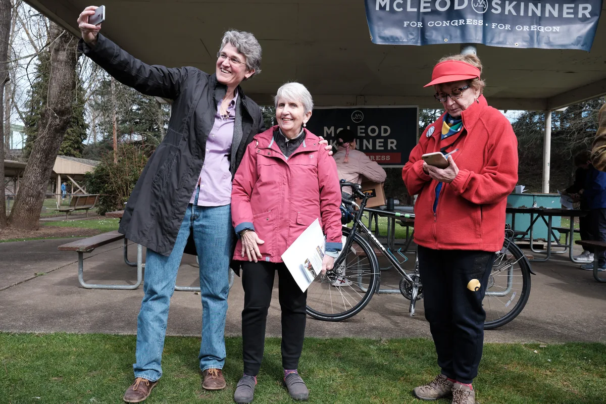 Three women at a political campaign event in Oregon City, with one woman raising her arm in celebration while standing next to campaign materials under a park pavilion. A McLeod Skinner for Congress banner is visible in the background along with bicycles and picnic tables.