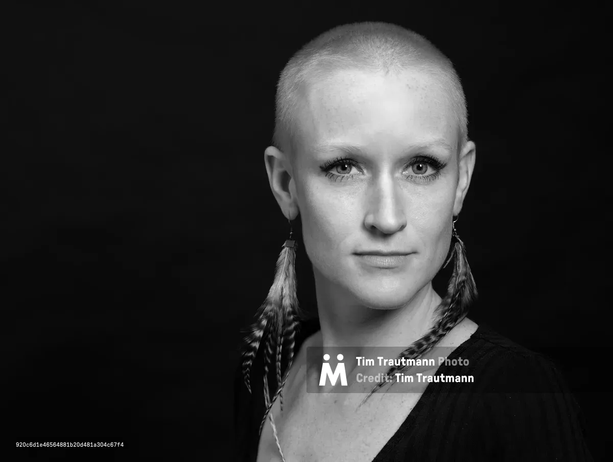 Cayce Ryan gazes directly into the camera with quiet intensity against a deep black backdrop in this striking monochrome headshot. Dramatic studio lighting sculpts her face with precision, creating subtle shadows that emphasize her defined features and cropped hair. Distinctive feather earrings catch the light, adding textural contrast to the minimalist composition. The professional portrait captures both vulnerability and strength, perfectly suited for her resume and social media presence.