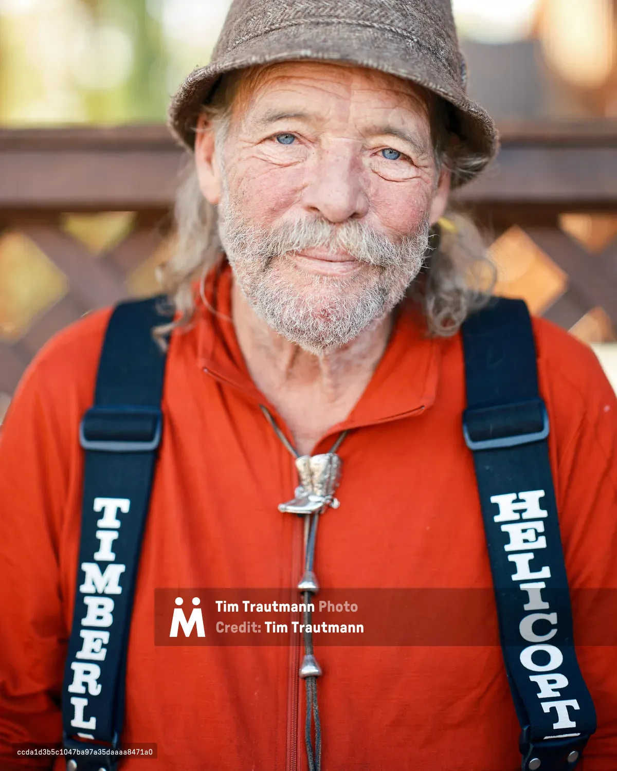A portrait of forester and poet Mark Miller in Spray, Oregon. He is an older man with blue eyes, a grey beard, and long grey hair, wearing a red zip-up fleece, a grey bucket hat, and a silver cowboy boot bolo tie. Black suspenders printed with the words "TIMBERLINE" and "HELICOPTERS" cross his chest. He smiles warmly at the camera, with a wooden lattice fence and soft bokeh foliage visible in the background.