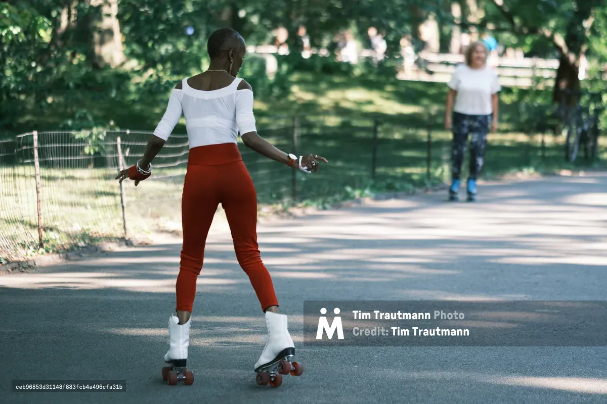 A woman glides gracefully on roller skates along a sun-dappled path in Central Park, her white off-shoulder top and vibrant red pants creating a striking silhouette against the verdant canopy. The golden afternoon light filters through the dense foliage, casting dancing shadows across the asphalt as park visitors enjoy the peaceful setting in the background. Her movement captures the effortless joy of urban recreation, while the shallow depth of field creates an intimate moment within the bustling heart of Manhattan.