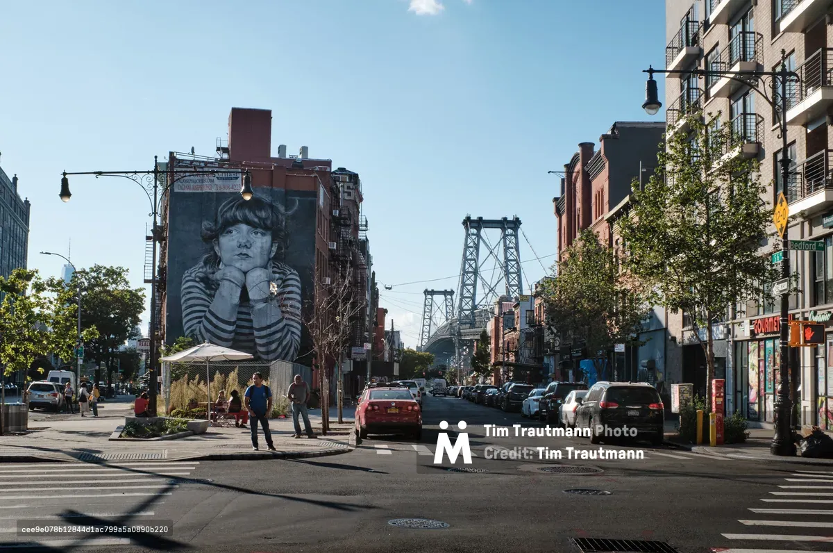 A striking black and white mural of a contemplative child dominates the brick facade of a building on Bedford Avenue in Williamsburg, Brooklyn. The monochromatic portrait creates a powerful contrast against the warm afternoon light that bathes the bustling street scene, where pedestrians cross the intersection as traffic flows toward the iconic Manhattan Bridge towers visible in the distance. The composition captures the vibrant street life of this gentrified neighborhood, where contemporary residential buildings with modern balconies stand alongside older brick structures adorned with large-scale street art.