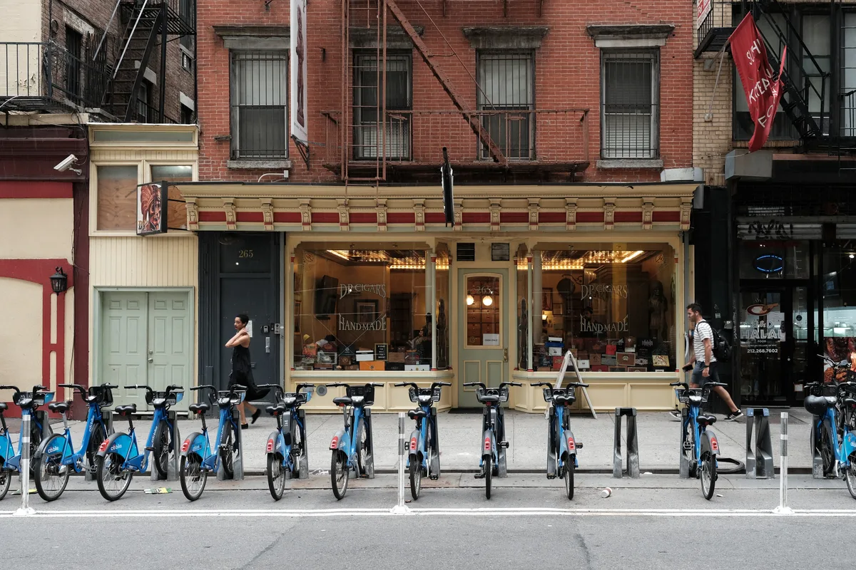 A traditional cigar shop with warm golden storefront lighting anchors this Chelsea street scene on 8th Avenue. The classic brick tenement facades create a rich tapestry of red and cream architectural details, while a neat row of blue Citi Bikes lines the sidewalk in perfect urban symmetry. Pedestrians move casually through the frame as late afternoon light bathes the handmade cigar shop's inviting windows, creating an intimate portrait of Manhattan neighborhood commerce.