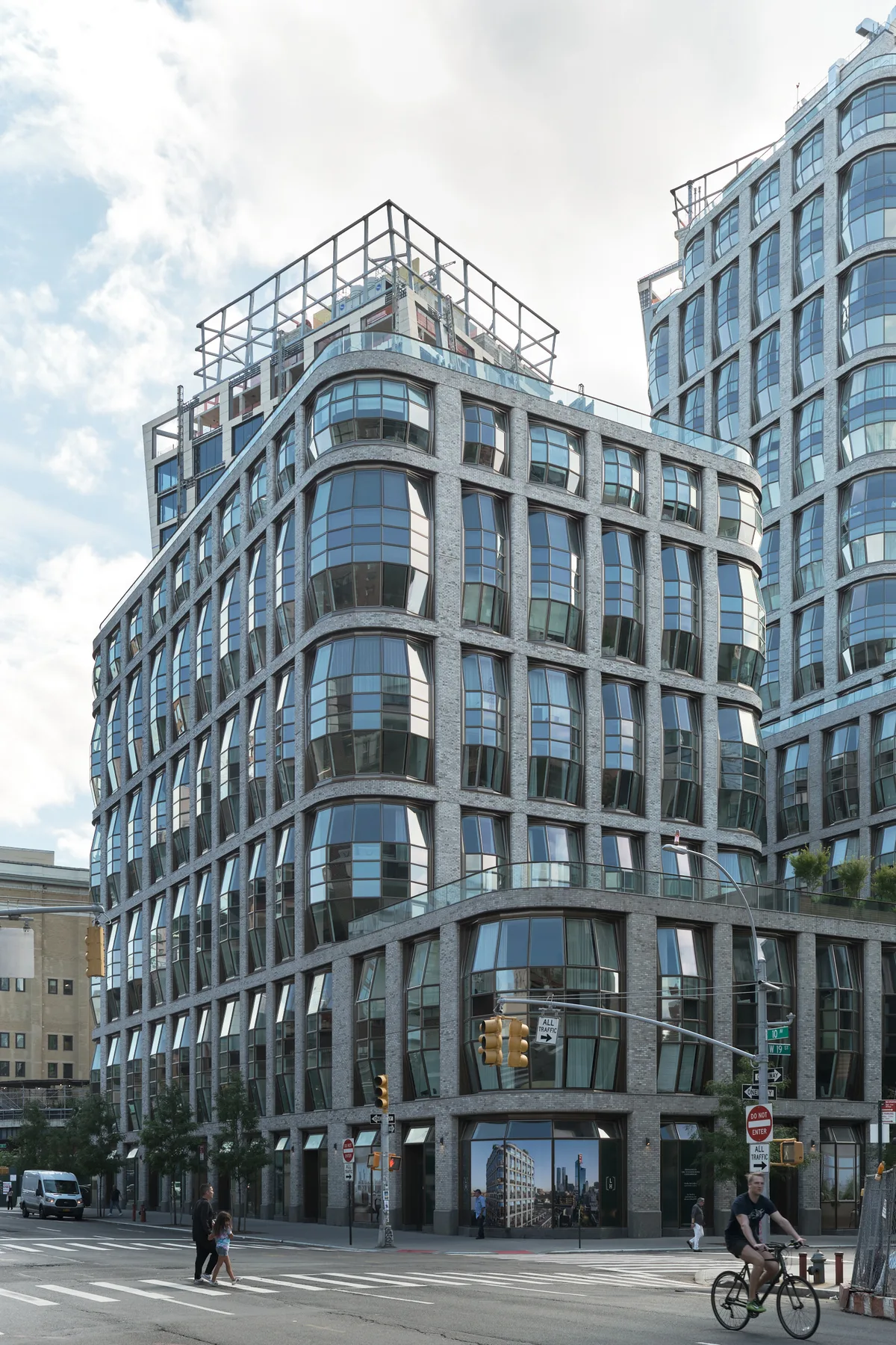 A dramatic upward perspective captures the sweeping curves of a contemporary residential tower on West 19th Street in Chelsea, its undulating glass facade creating a rhythmic pattern against overcast skies. The building's distinctive rounded corner anchors the intersection, while pedestrians and a cyclist navigate the crosswalk below, their small figures emphasizing the structure's imposing scale. Soft, diffused daylight illuminates the blue-tinted glass panels and gray stone framework, creating subtle reflections that dance across the building's sinuous surface.