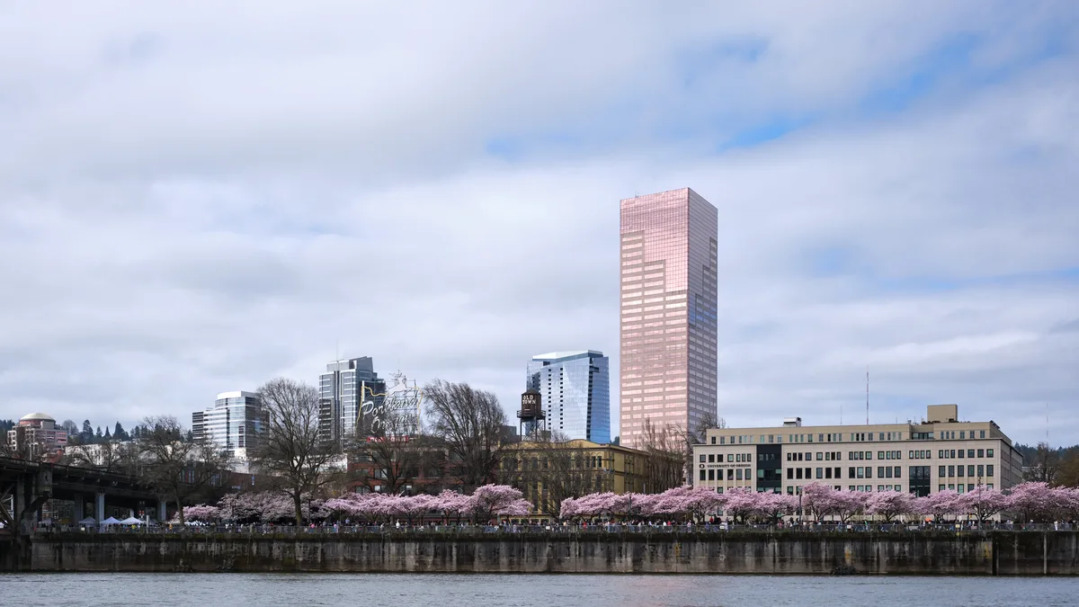 Delicate pink cherry blossoms create a romantic foreground along the Willamette River waterfront, contrasting beautifully with Portland's modern downtown skyline. The iconic pink tower rises majestically above the historic brick buildings and contemporary glass structures, while visitors stroll beneath the blooming canopy at Tom McCall Waterfront Park. Soft, diffused light filters through the overcast sky, creating a dreamy atmosphere that captures the ephemeral beauty of spring in the Pacific Northwest.