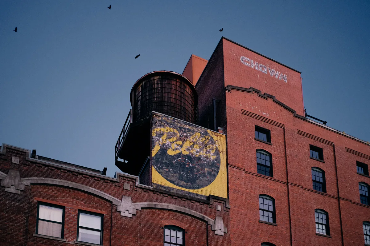A low-angle view of the historic Chown Pella building in Portland, Oregon, photographed at dusk against a deep blue sky. The multi-story red brick industrial building features a vintage wooden water tower on its roof and a large weathered painted advertisement reading "Pella" in yellow script on a circular dark background. The faded "Chown" name is painted in ghost lettering on the upper brick facade. Several birds are silhouetted against the darkening sky above the roofline.