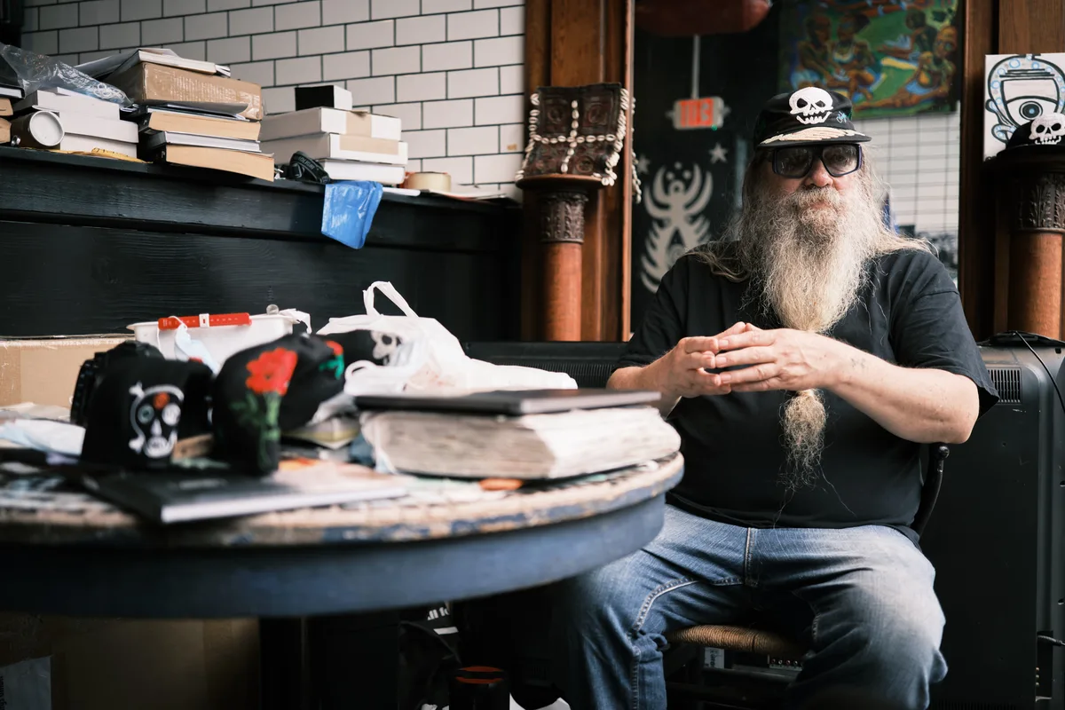 Folk historian and documentarian Clayton Patterson sits contemplatively in his cluttered Essex Street studio, his distinctive long gray beard and skull-adorned cap reflecting decades of chronicling Manhattan's Lower East Side. Warm afternoon light filters through the space, illuminating stacks of books, scattered baseball caps with skull motifs, and the accumulated detritus of a life spent preserving neighborhood history. His weathered hands rest thoughtfully as he surveys his domain—part archive, part sanctuary—where the counter-cultural spirit of old New York endures.