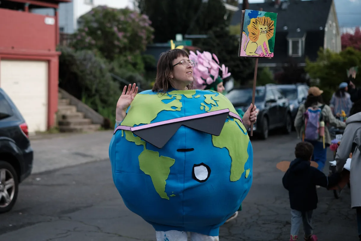 A person wearing a large spherical Earth costume with blue oceans and green continents stands on a Portland street, holding a protest sign while participating in what appears to be a climate change demonstration. The costume features sunglasses and the person is waving at the camera.