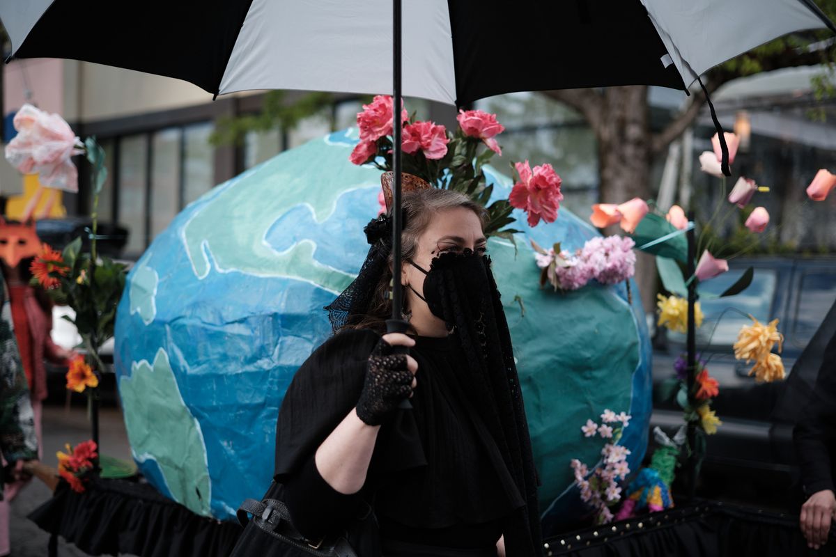 A person wearing all black with a face mask stands under a black and white umbrella, adorned with a large blue and green Earth costume decorated with colorful flowers. The scene appears to be at an outdoor environmental protest or demonstration in Portland, Oregon.