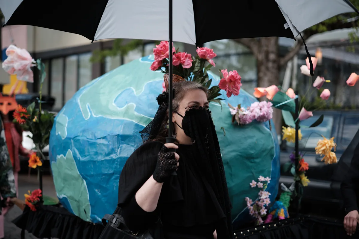 A person wearing all black with a face mask stands under a black and white umbrella, adorned with a large blue and green Earth costume decorated with colorful flowers. The scene appears to be at an outdoor environmental protest or demonstration in Portland, Oregon.