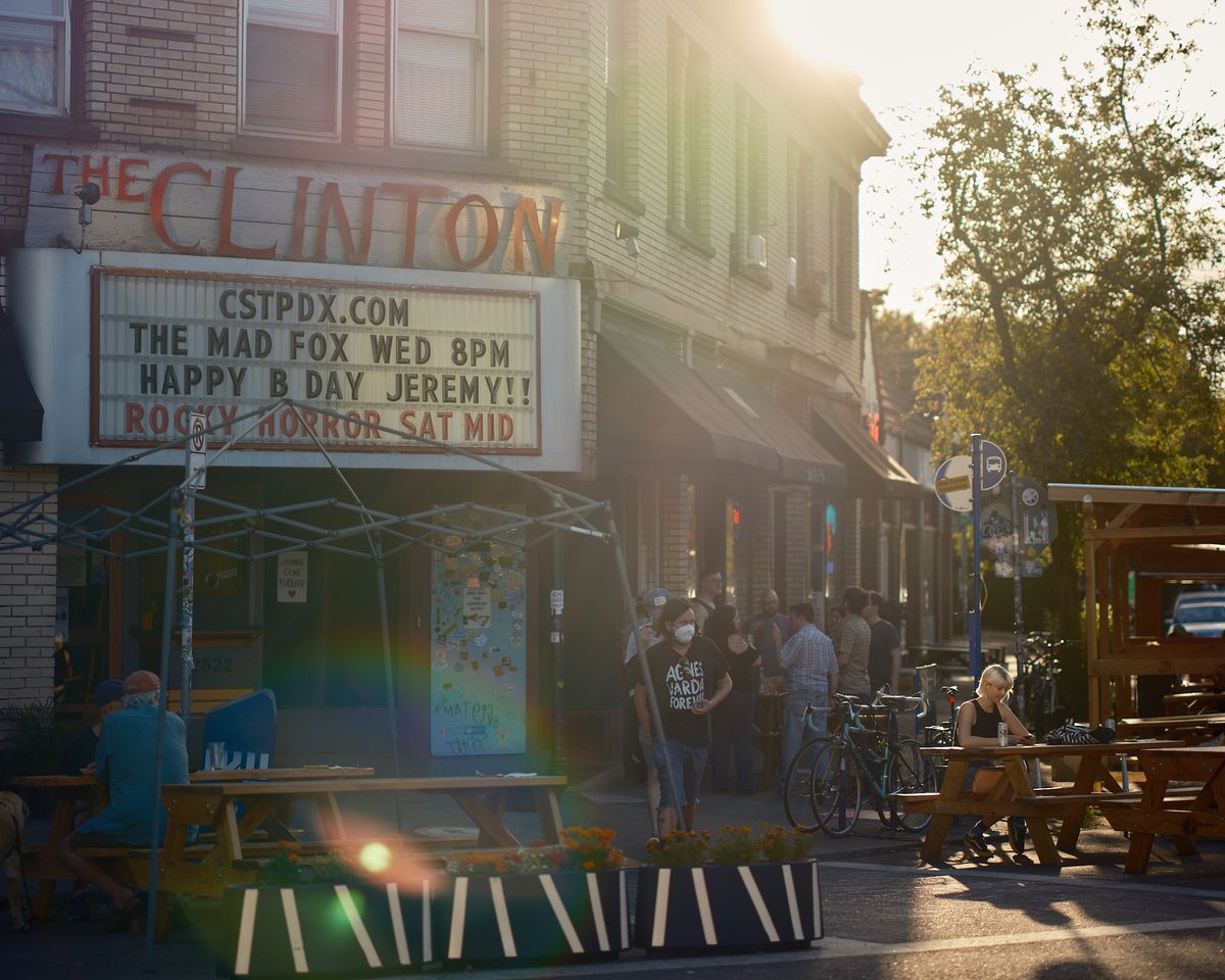 Alt text:  The exterior of The Clinton Street Theater in Portland, Oregon, bathed in warm golden lens-flare sunlight. The theater's marquee reads "The Mad Fox Wed 8PM — Happy B Day Jeremy!! — Rocky Horror Sat Mid." A lively crowd of people gathers on the sidewalk outside, with outdoor picnic tables and planters with marigolds occupying a parklet in the foreground. A woman in a face mask wearing a black "Agnès Varda Forever" t-shirt walks past. Bicycles are parked along the curb, and a woman sits alone at a picnic table to the right.