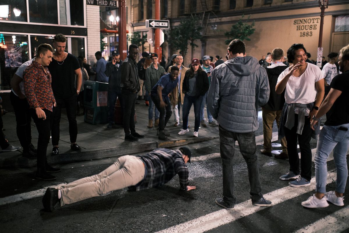 A late-night street scene at the corner of NW Third Avenue in Portland's Chinatown as bars and clubs close for the night. A young man in a plaid shirt and khaki pants does push-ups in the middle of the wet street while a crowd of onlookers gathers around him, some laughing and watching with amusement. The historic Sinnott House building from 1883 is visible in the background, along with a one-way street sign and the glow of bar lights on the rain-slicked sidewalk.