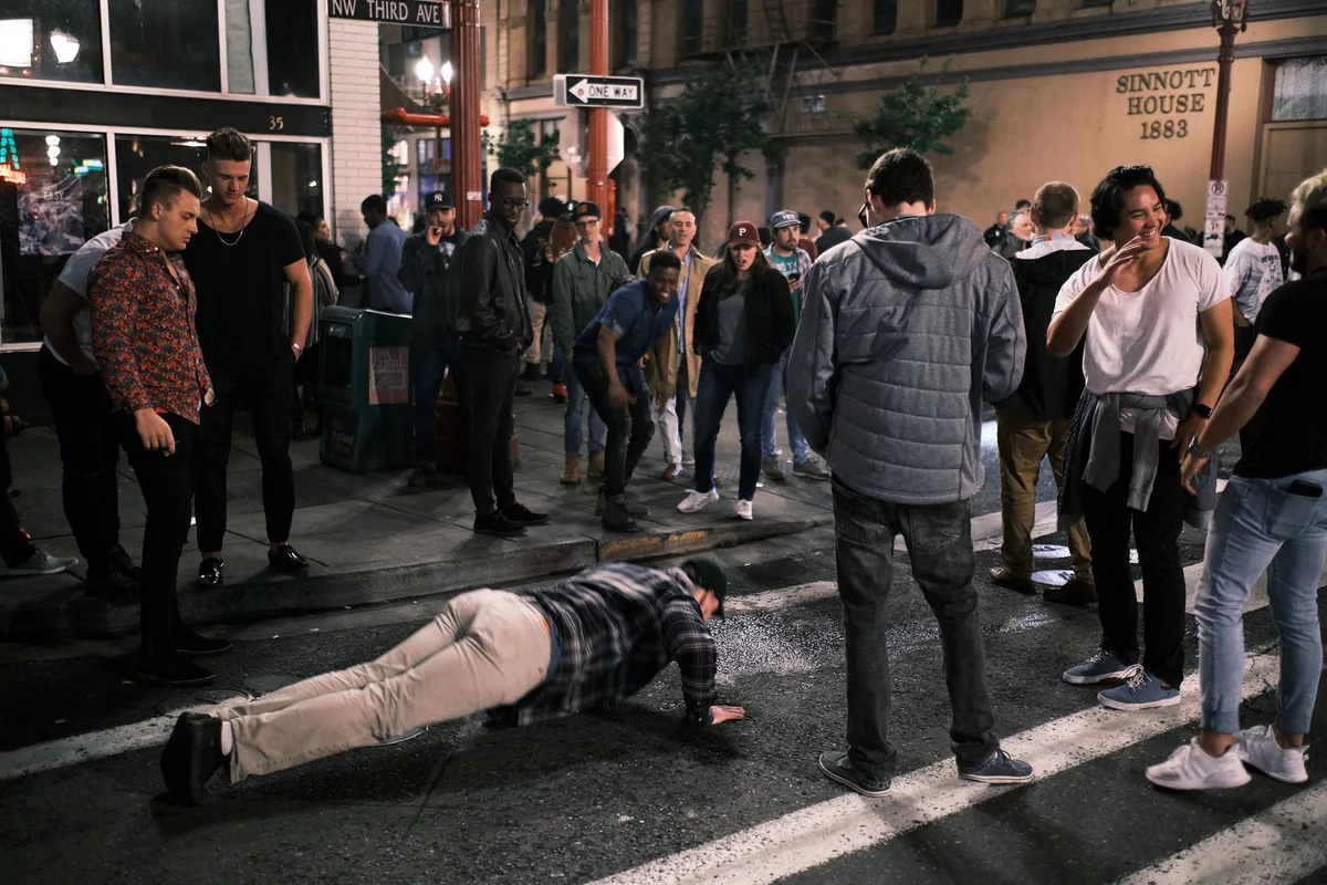 A late-night street scene at the corner of NW Third Avenue in Portland's Chinatown as bars and clubs close for the night. A young man in a plaid shirt and khaki pants does push-ups in the middle of the wet street while a crowd of onlookers gathers around him, some laughing and watching with amusement. The historic Sinnott House building from 1883 is visible in the background, along with a one-way street sign and the glow of bar lights on the rain-slicked sidewalk.