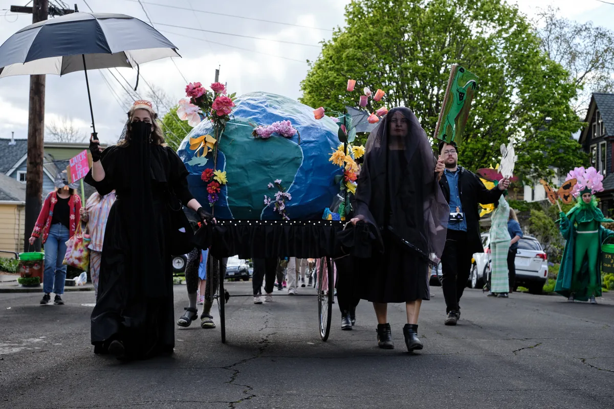 A group of environmental activists dressed in black participate in a community parade in Portland, Oregon, carrying a large Earth globe decorated with flowers on a wheeled cart while walking down a residential street lined with trees and houses.