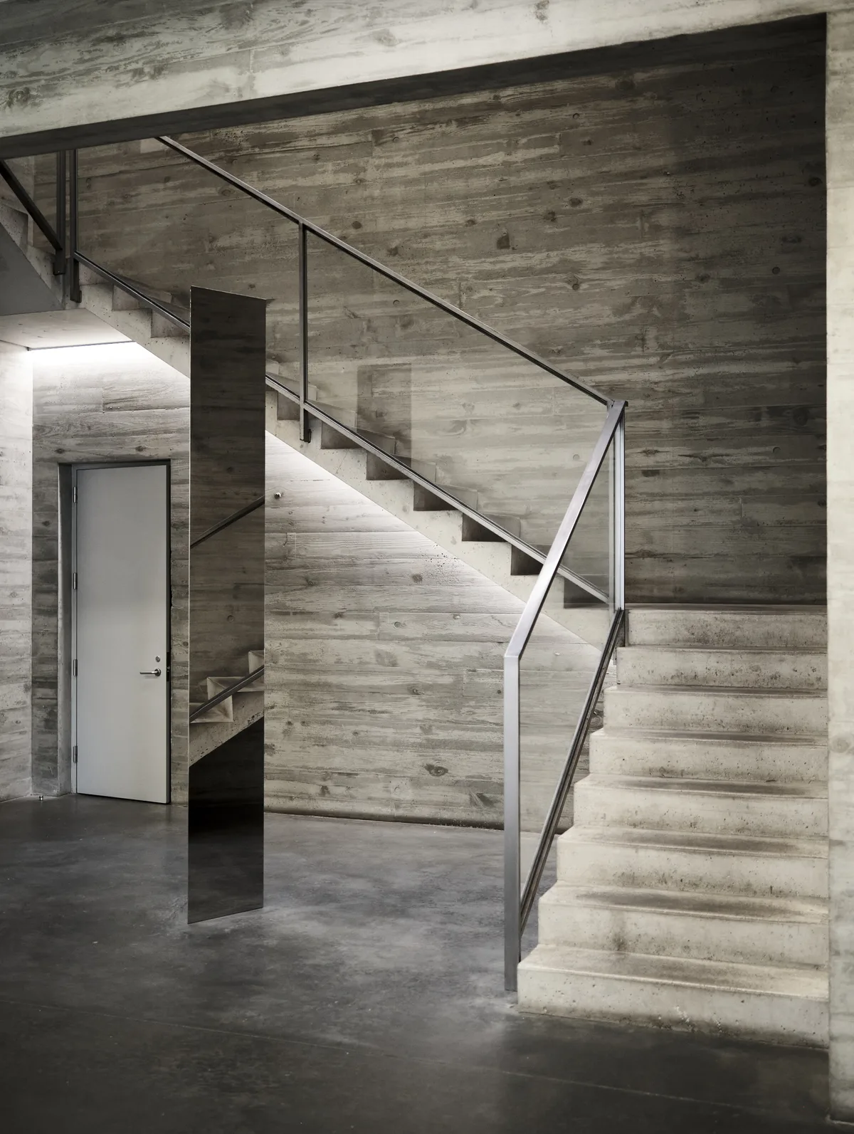 A monumental concrete staircase ascends through raw architectural space, its board-formed surfaces bearing the ghostly imprints of wooden planks. Steel handrails trace geometric lines against weathered concrete walls, while muted natural light filters through the brutalist interior. The composition emphasizes the interplay between rough-hewn materiality and precise modern engineering, creating a cathedral-like atmosphere within this Chelsea arts district building.