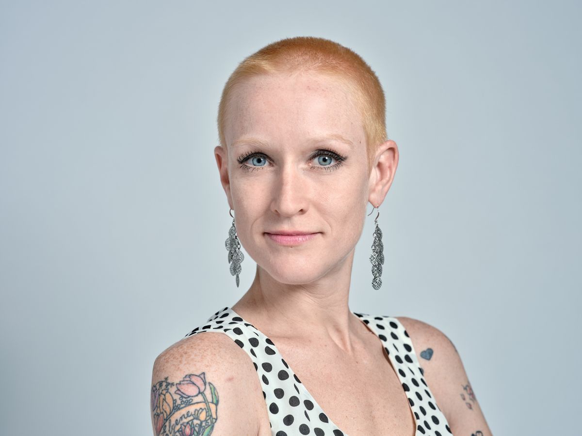 Cayce Ryan presents a confident, contemporary portrait against a clean studio backdrop in Portland's industrial Southeast district. Her platinum buzz cut frames piercing blue eyes enhanced with subtle makeup, while vintage-inspired drop earrings complement her polka-dotted halter top. Colorful tattoo artwork adorns her shoulders, creating visual interest against the minimalist composition and soft, even lighting that emphasizes her professional yet artistic persona.