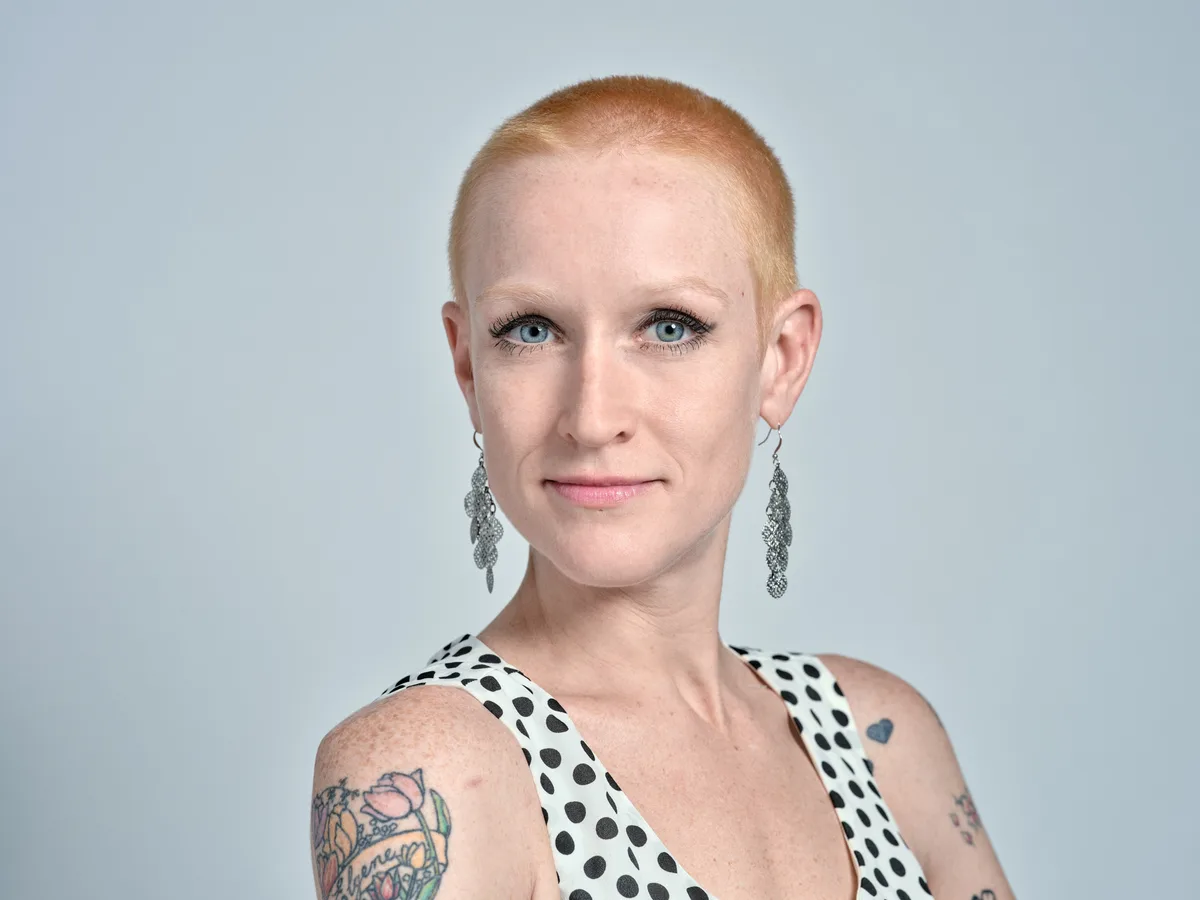 Cayce Ryan presents a confident, contemporary portrait against a clean studio backdrop in Portland's industrial Southeast district. Her platinum buzz cut frames piercing blue eyes enhanced with subtle makeup, while vintage-inspired drop earrings complement her polka-dotted halter top. Colorful tattoo artwork adorns her shoulders, creating visual interest against the minimalist composition and soft, even lighting that emphasizes her professional yet artistic persona.