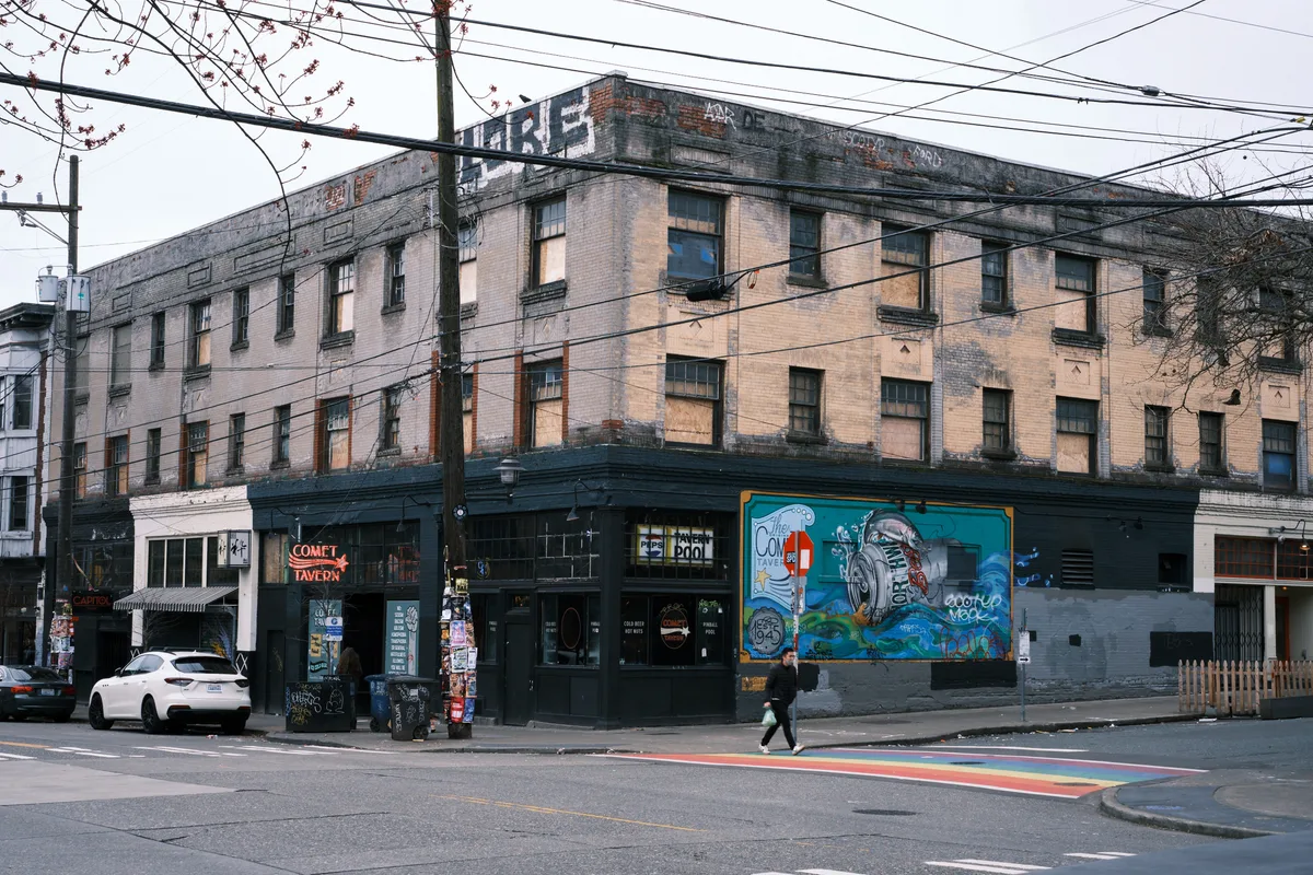 The Comet Tavern anchors a weathered brick corner building in Seattle's Capitol Hill neighborhood, where power lines slice through an overcast sky above rain-slicked streets. The building's faded industrial facade tells stories of urban evolution, with peeling paint and graffiti creating a textural palimpsest against warm brick tones. A solitary figure crosses the intersection marked by rainbow crosswalk stripes, while vintage neon signs and vibrant street murals inject color into the moody Pacific Northwest atmosphere.