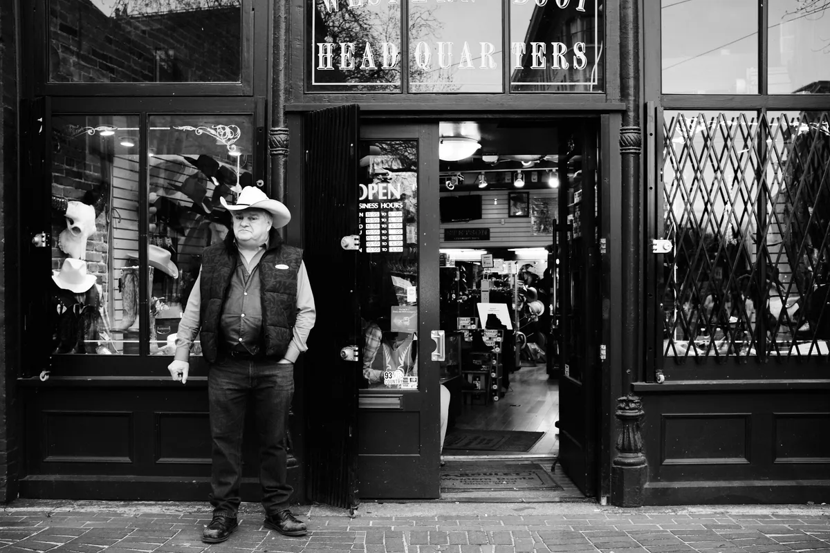 Tony Chisholm, a sales associate at OK Boot Corral, takes a contemplative break outside the Western wear store in Vancouver's historic Gastown district. Dressed in authentic cowboy attire—white Stetson hat, dark vest, and weathered boots—he leans casually against the ornate storefront of Byrnes Block on Carrall Street. The black and white composition captures the timeless intersection of frontier heritage and urban commerce, with vintage display windows revealing leather goods and Western memorabilia within.