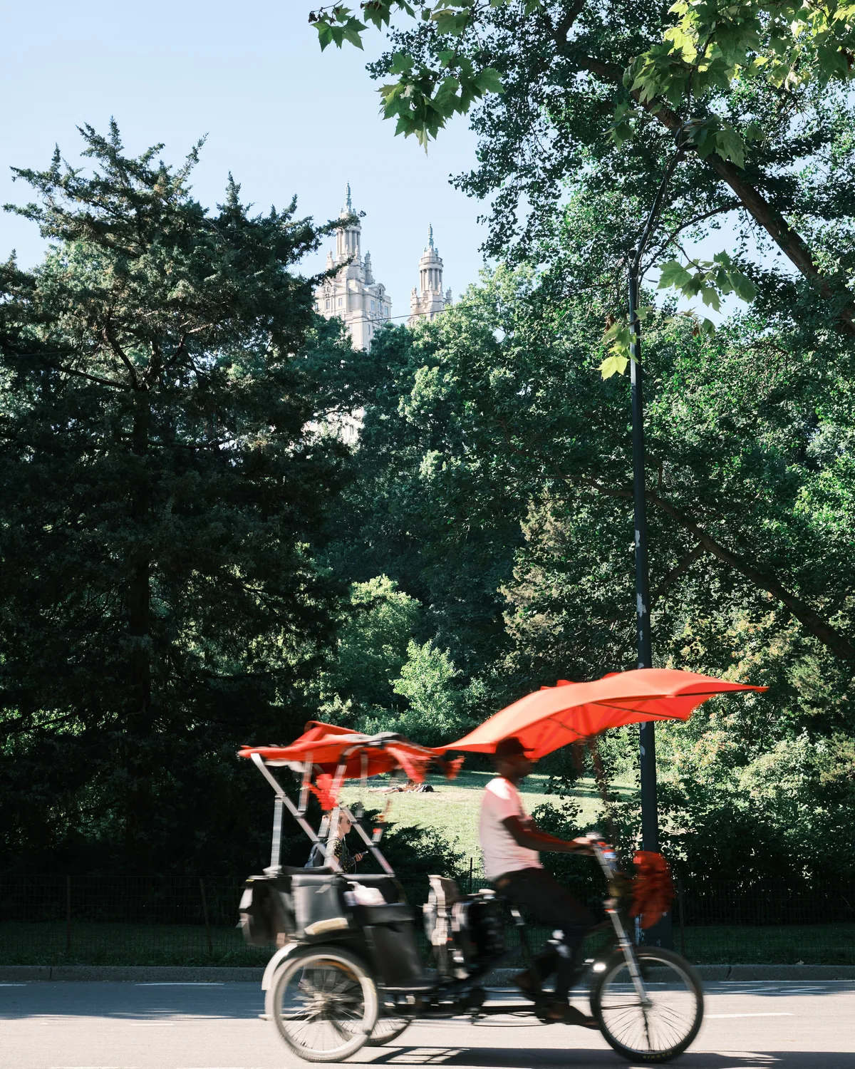 A pedicab adorned with billowing vermillion fabric cuts through dappled sunlight beneath Central Park's dense canopy, its motion-blurred form suggesting the unhurried rhythm of urban leisure. The Gothic Revival spires of the San Remo apartments pierce the green horizon beyond, their limestone towers creating a romantic backdrop to this fleeting moment of transportation. Filtered golden light cascades through maple and oak leaves, casting the scene in the warm, nostalgic glow of a perfect New York summer afternoon.