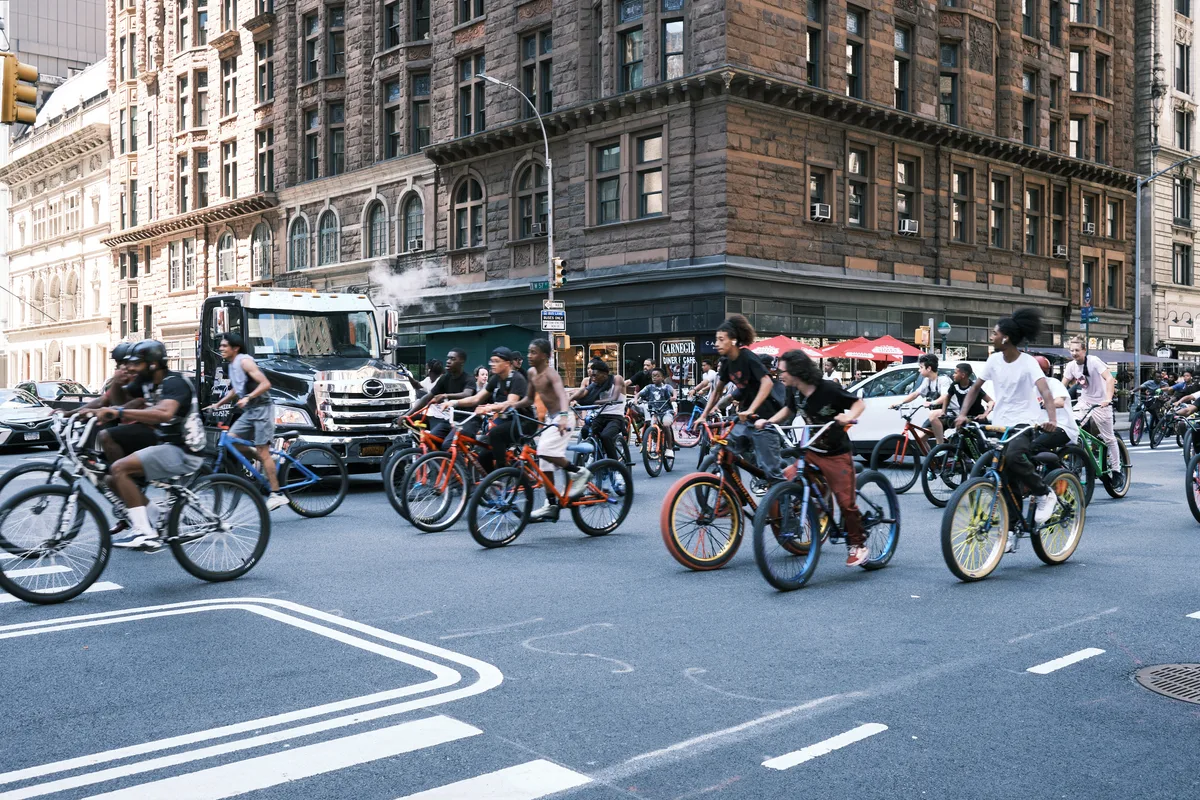 A dynamic river of cyclists flows through the intersection at 7th Avenue in Midtown Manhattan, their colorful bicycles creating streaks of motion against the backdrop of historic brownstone architecture. The late afternoon light bathes the weathered facades in warm golden tones while participants in what appears to be a Critical Mass ride reclaim the urban streetscape. Motion blur captures the kinetic energy of dozens of riders as they navigate the intersection, transforming the typically car-dominated thoroughfare into a temporary cycling sanctuary.