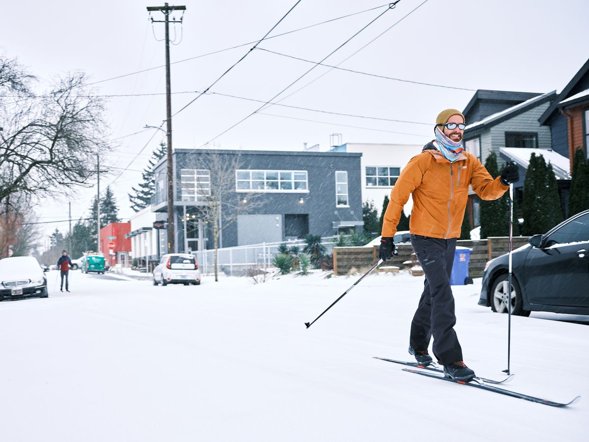 A man cross-country skis down a snow-covered residential street in Portland's Alberta Arts District during a winter snowstorm. He wears an orange jacket, black ski pants, a mustard yellow beanie, and mirrored sunglasses, smiling as he glides past parked cars. A second skier in a red jacket is visible further down the street in the background, along with modern homes, utility poles, and lightly falling snow.