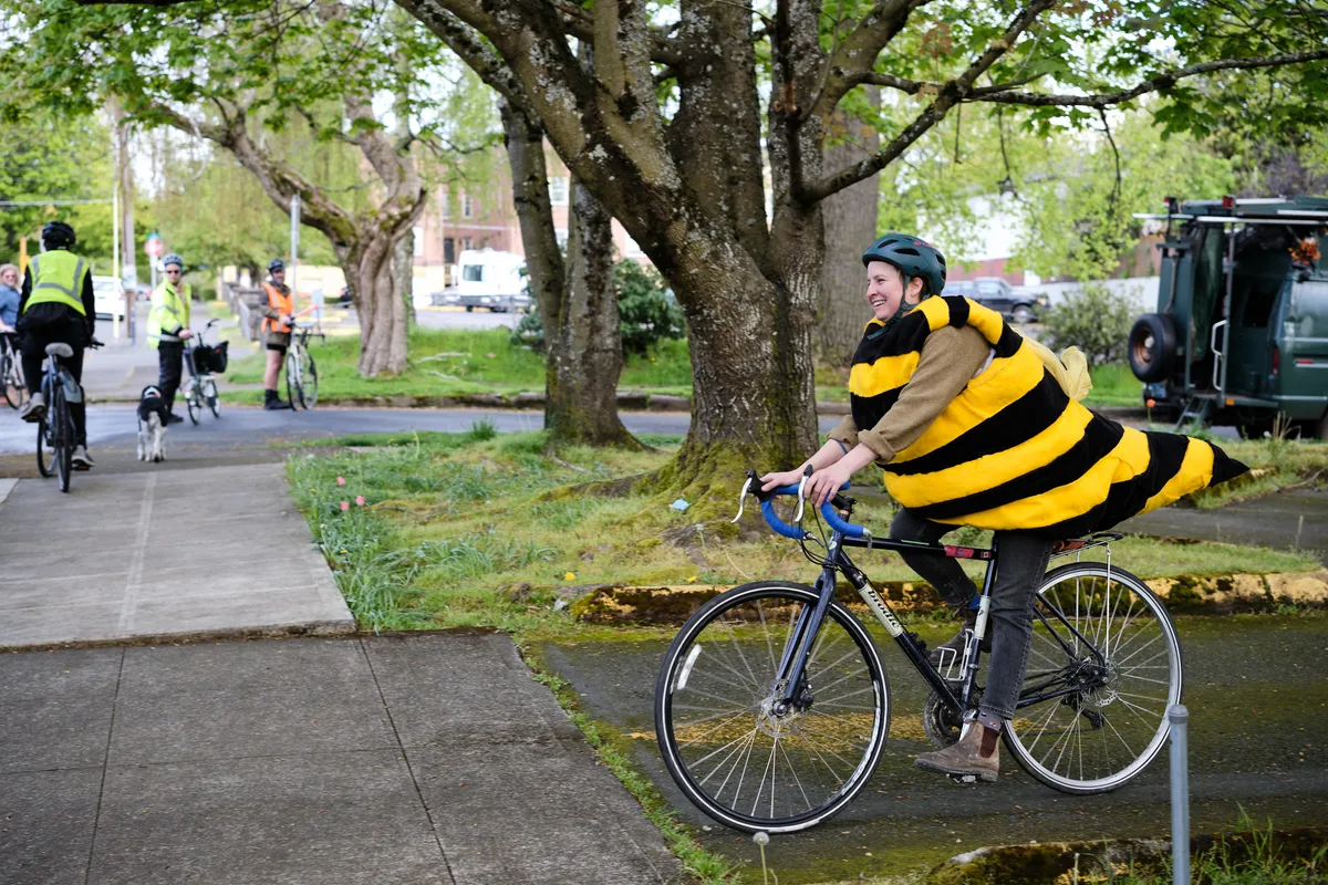 A smiling cyclist wearing a yellow and black striped bee costume rides a bicycle on a tree-lined street in Portland's Buckman neighborhood. Other cyclists in safety vests can be seen in the background, suggesting this is part of a community cycling event or parade.