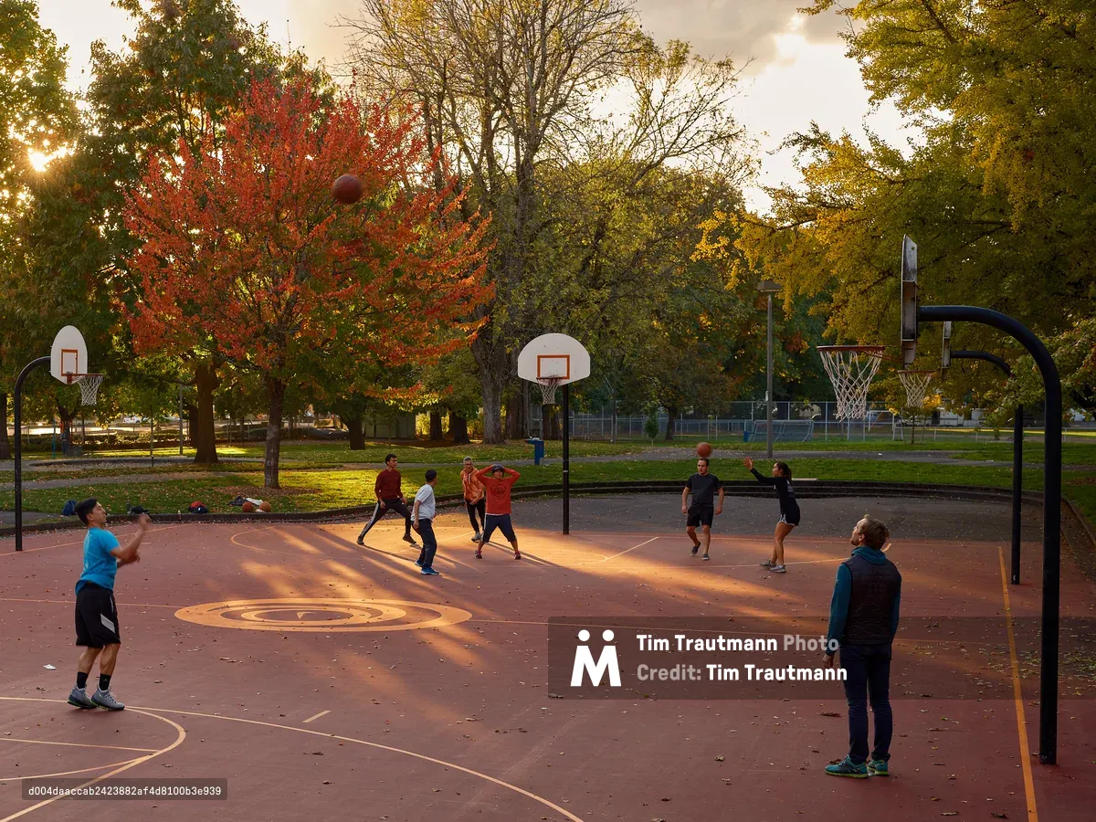 A group of people play pickup basketball on an outdoor court at Powell Park in Portland, Oregon, bathed in warm golden evening light. A basketball is suspended mid-air above the court. Three hoops are visible across the wide red-surfaced court, with fallen autumn leaves scattered across it. Vibrant fall foliage in shades of red, orange, and yellow fills the background, with the setting sun breaking through the trees on the left.
