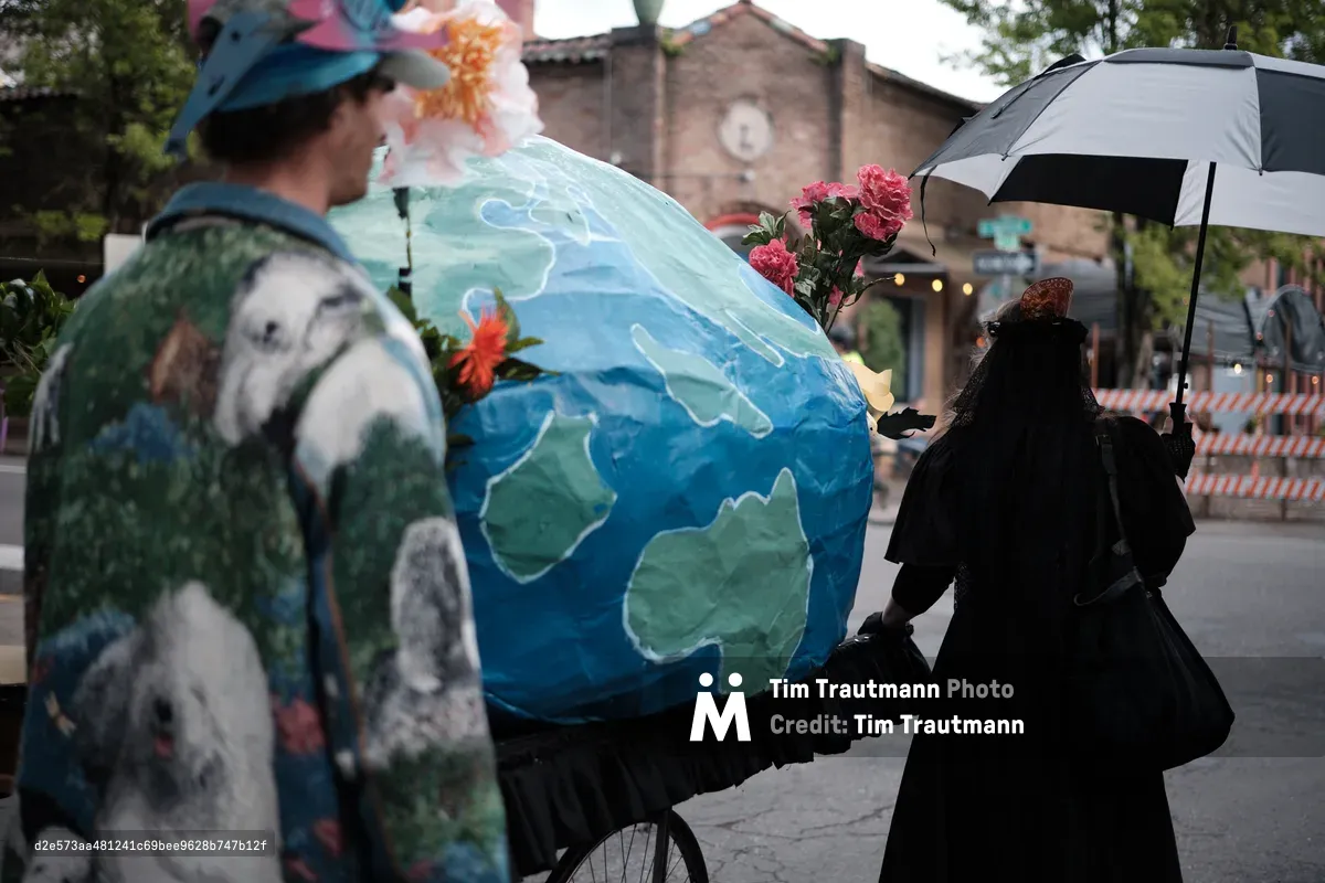 A street performance scene in Portland's Central Eastside district featuring performers with a large painted Earth globe prop decorated with flowers, while a person in dark clothing holds an umbrella nearby. The urban setting includes brick buildings and trees in the background.