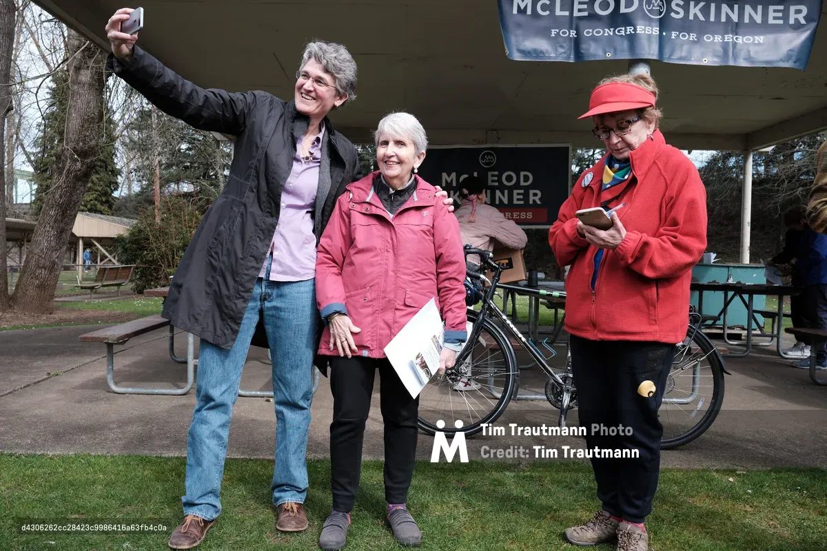 Three women at a political campaign event in Oregon City, with one woman raising her arm in celebration while standing next to campaign materials under a park pavilion. A McLeod Skinner for Congress banner is visible in the background along with bicycles and picnic tables.