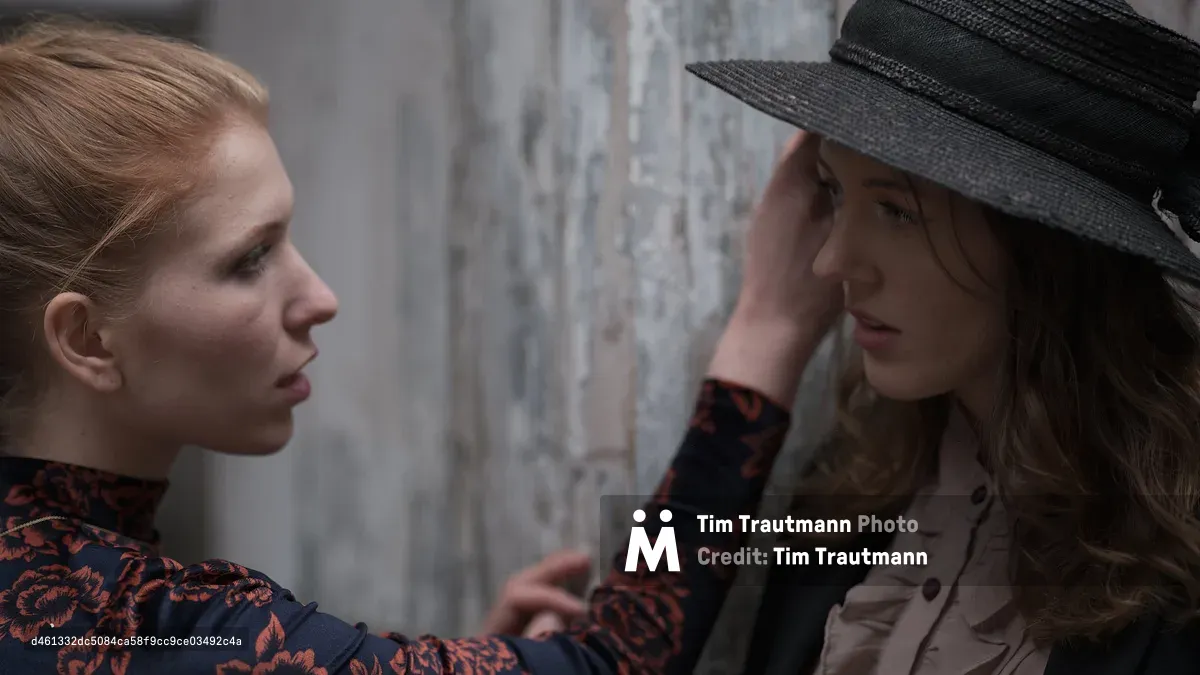 A close, intimate portrait of two women facing each other against a weathered concrete wall. Faith Yakovleva, seen in profile on the left, wears a dark floral top with red and black flowers and has her strawberry blonde hair pulled back. She reaches out to gently touch the brim of a wide black straw hat worn by Rhianna Atwood, who stands against the wall on the right in a cream ruffled blouse, her eyes downcast.