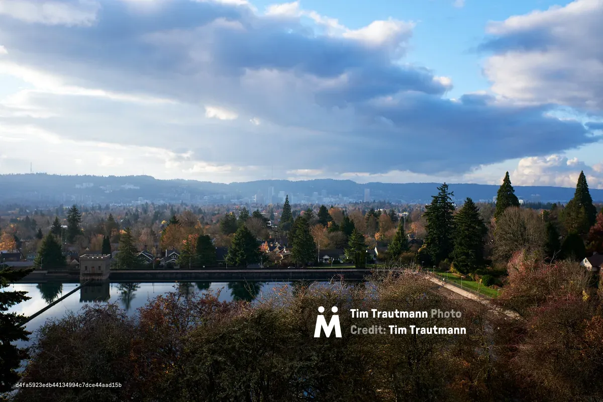 Storm clouds gather dramatically over Portland's residential sprawl as viewed from Mount Tabor's reservoir overlook during peak autumn foliage. The serene water surface mirrors the moody sky while evergreen sentinels frame the foreground, their dark forms contrasting against the warm amber and rust tones of deciduous trees below. The city extends toward distant hills under brooding cumulus formations, creating a contemplative tableau that captures the Pacific Northwest's characteristic interplay between urban development and natural beauty.