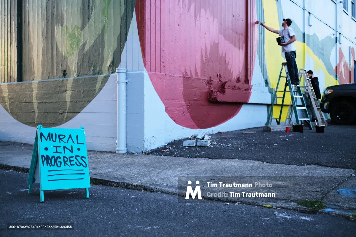 An artist perched on a ladder applies bold strokes to a sweeping geometric mural adorning the exterior wall of Coast Auto Supply on Southeast Stark Street. The composition features flowing bands of sage green, dusty rose, and sunshine yellow that curve across the weathered concrete surface. A cheerful turquoise sidewalk sign reading 'MURAL IN PROGRESS' sits on the cracked asphalt, while afternoon light catches the fresh paint and casts long shadows across the urban streetscape.