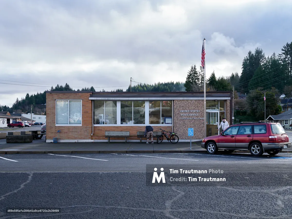 A modest brick and stone United States Post Office sits beneath brooding clouds in South Bend, Washington, embodying the quiet functionality of small-town America. The mid-century modern building, with its expansive windows and clean lines, anchors this Pacific County streetscape where a burgundy SUV idles alongside patrons conducting their daily postal business. Dense evergreen forests form a verdant backdrop against the overcast sky, while the American flag stands sentinel above this essential community hub. The scene captures the unhurried rhythm of rural life in the Pacific Northwest, where civic architecture serves as both practical necessity and social gathering point.