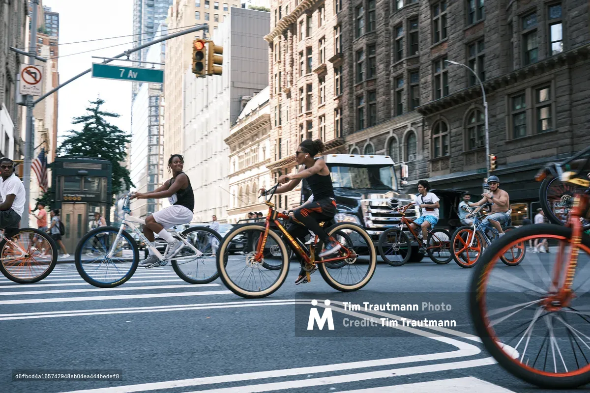 A dynamic street scene captures cyclists in motion along 7th Avenue in Manhattan's Midtown, their wheels blurred against the crisp asphalt crosswalk stripes. The golden hour light bathes the historic stone facades and modern glass towers that define this iconic corridor, while pedestrians and vehicles share the urban rhythm. The composition emphasizes the democratic nature of city streets, where bicycles weave through the metropolitan tapestry alongside cars and buses.