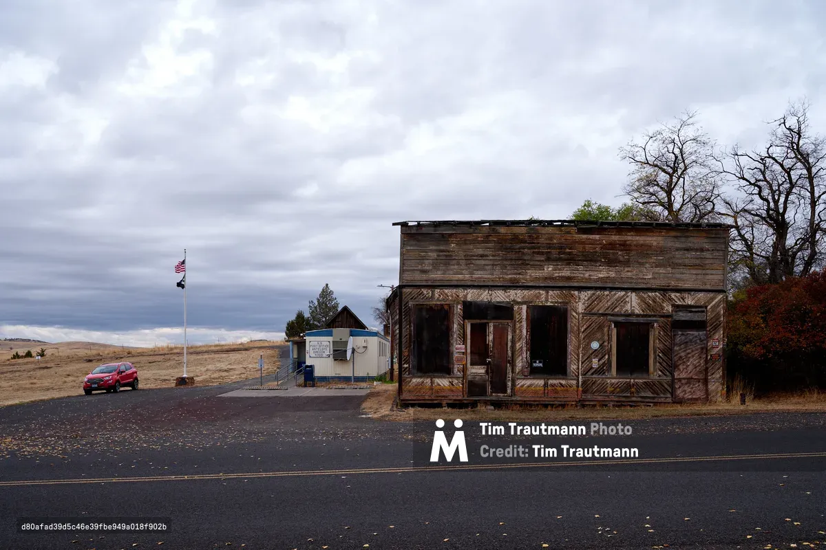 A weathered wooden storefront stands sentinel along a lonely stretch of Oregon highway, its diagonal siding bleached and buckled by high desert winds. The abandoned building, once Antelope's commercial heart, faces a small USPS trailer that now serves the scattered community of fewer than fifty souls. Under brooding November clouds, a red SUV idles beside the American flag, creating a stark tableau of rural isolation in Wasco County's vast wheat country. Bare cottonwoods frame the scene, their skeletal branches echoing the building's own slow surrender to time and elements.