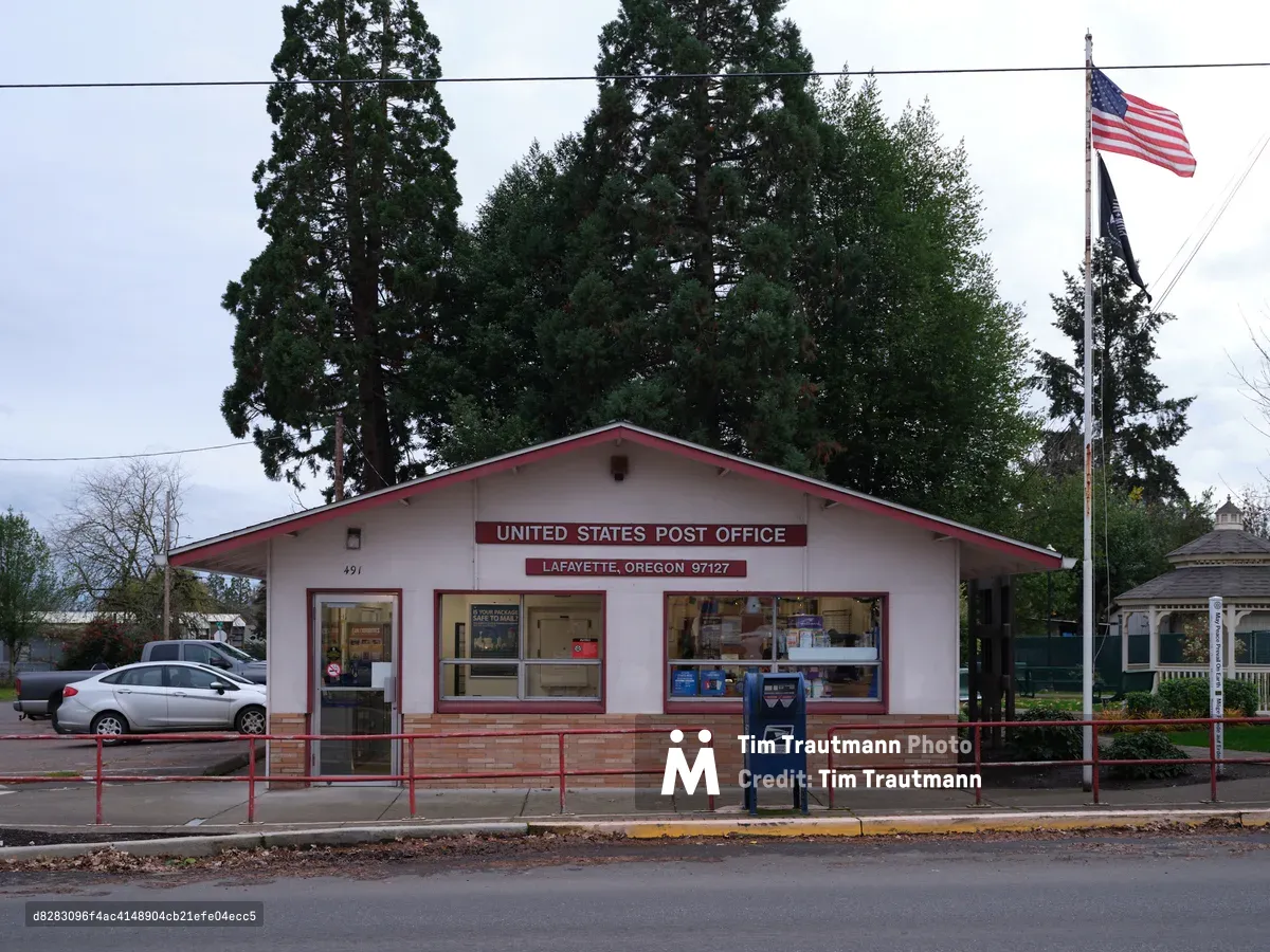 A modest single-story post office building stands beneath towering evergreens in the small Oregon town of Lafayette, its cream-colored walls accented with burgundy trim and signage. The American flag waves proudly from a tall pole beside the building, while power lines stretch across an overcast sky that speaks to the Pacific Northwest's characteristic atmosphere. A blue USPS mailbox sits sentinel on the sidewalk, and cars parked nearby suggest the steady rhythm of daily community life in this rural Yamhill County settlement.