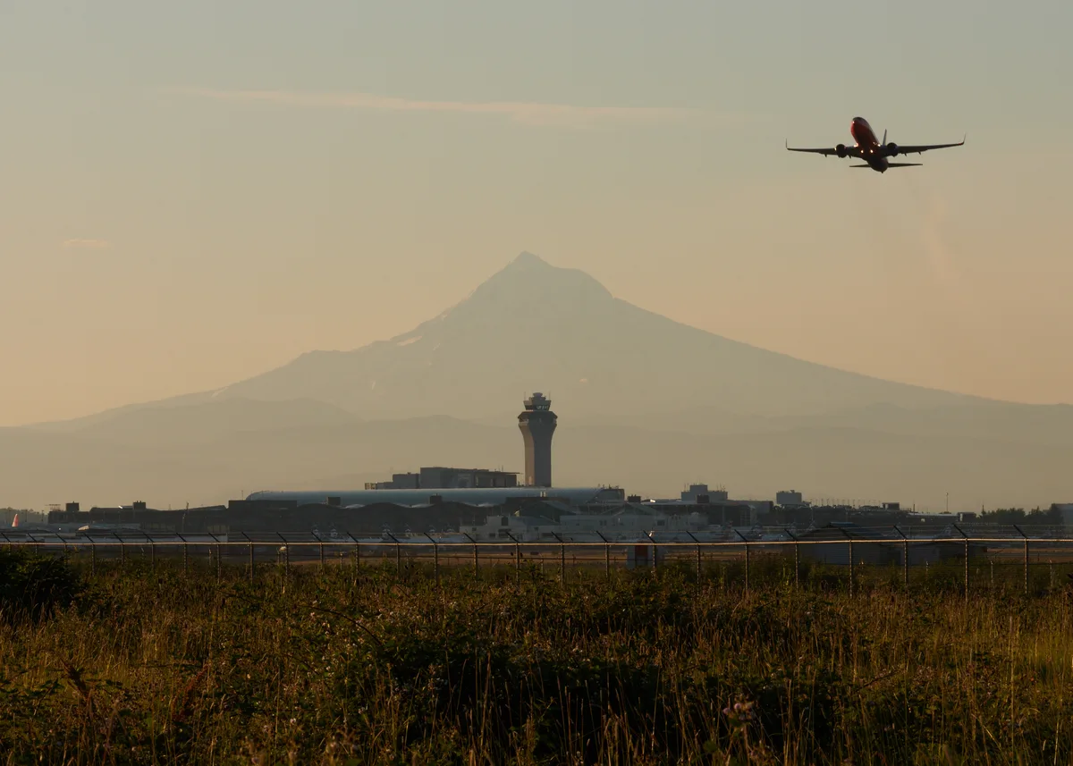 A commercial airliner climbs into the amber-hued twilight sky above Portland International Airport, silhouetted against the majestic volcanic cone of Mount Hood. The airport's distinctive control tower stands sentinel in the middle distance, while wild grasses in the foreground frame this layered composition of human aviation intersecting with Oregon's iconic Cascade peak. The warm, diffused light suggests either early morning or evening departure, casting the entire scene in a nostalgic, golden atmosphere.