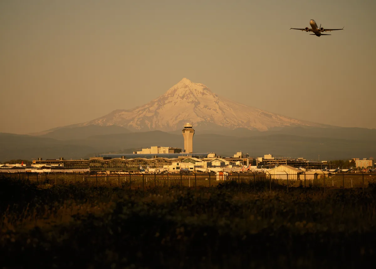A commercial aircraft ascends into the amber-washed sky above Portland International Airport, silhouetted against the imposing snow-capped peak of Mount Hood. The golden hour light bathes the airport's control tower and terminal buildings in warm honey tones, creating a striking contrast between human aviation infrastructure and Oregon's volcanic sentinel. Captured from the Sunderland neighborhood, this image transforms a routine departure into a cinematic moment where technology meets wilderness.