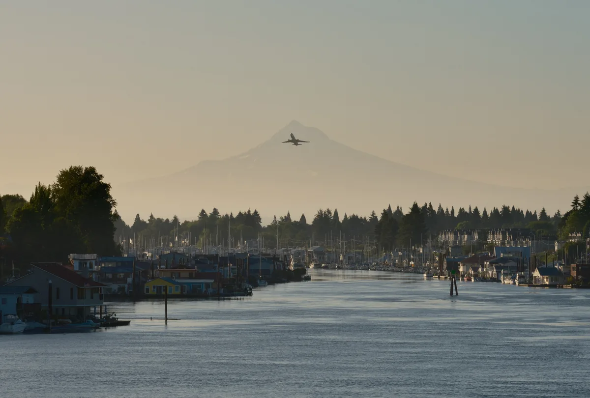 A commercial aircraft cuts through the pale morning sky above Mount Hood's ghostly silhouette, while the Columbia River winds past Portland's North Harbor in the golden hour light. The waterway reflects the muted tones of dawn as fishing boats and houseboats line the quiet marina, their masts creating a delicate forest against the distant Cascade Range. This serene moment captures the intersection of urban aviation and maritime life, where the rhythms of river commerce play out beneath the watchful presence of Oregon's iconic peak.